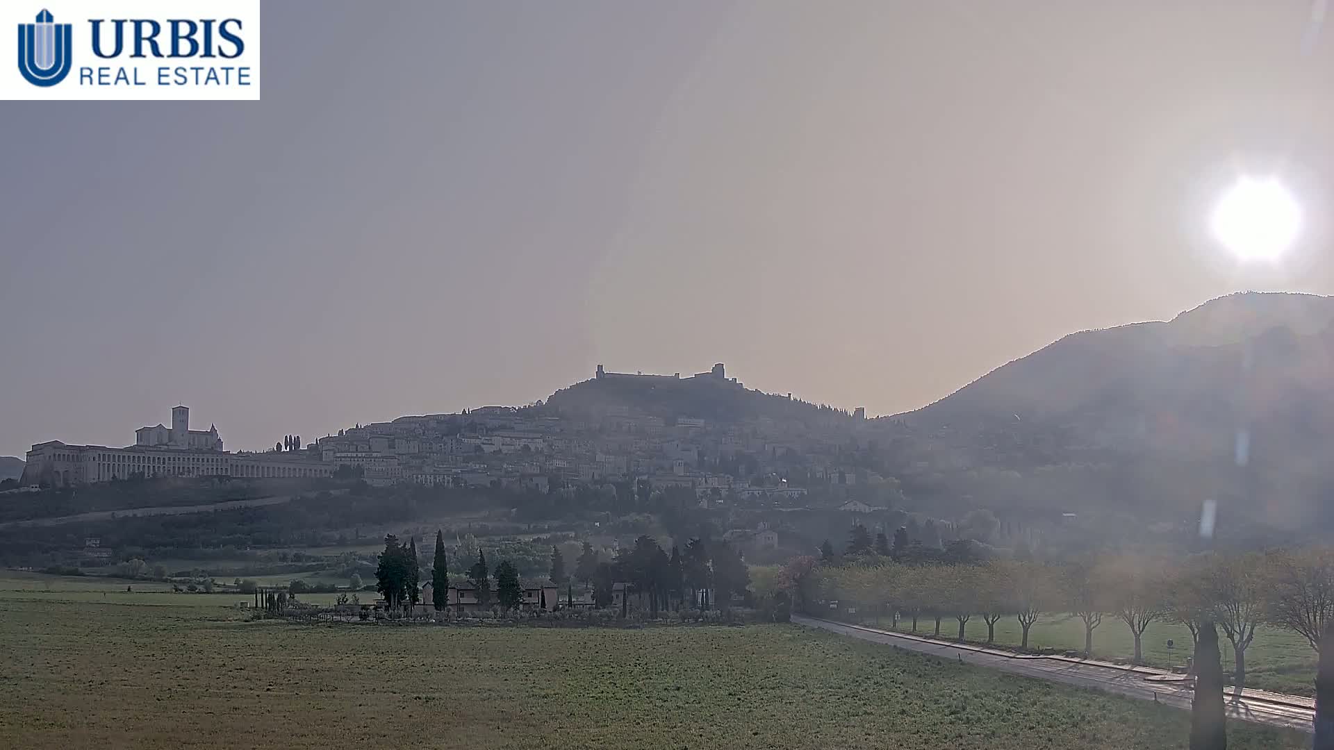 A hazy sunrise illuminates a hilltop town and fortress, with a flat, grassy foreground and a road lined with trees.