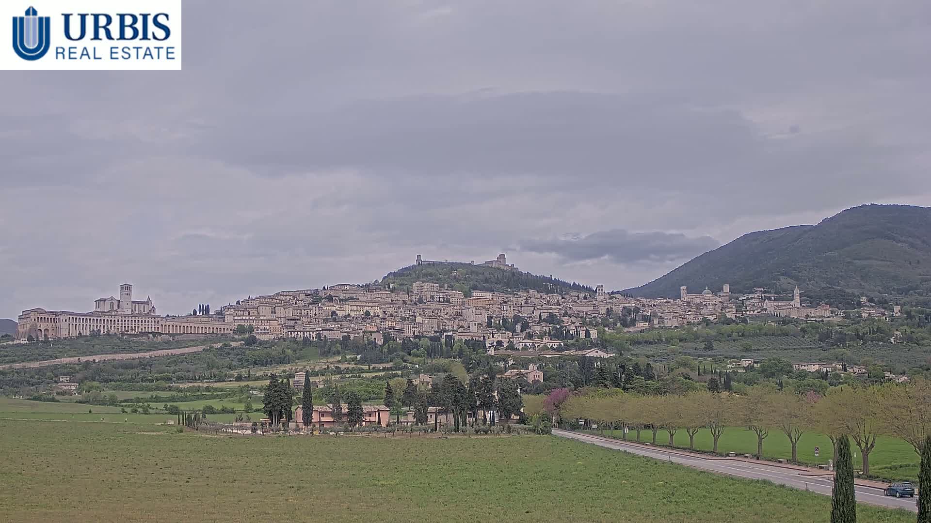 A hilltop town with numerous buildings, including a large basilica, is visible under an overcast sky, with green fields and trees in the foreground.