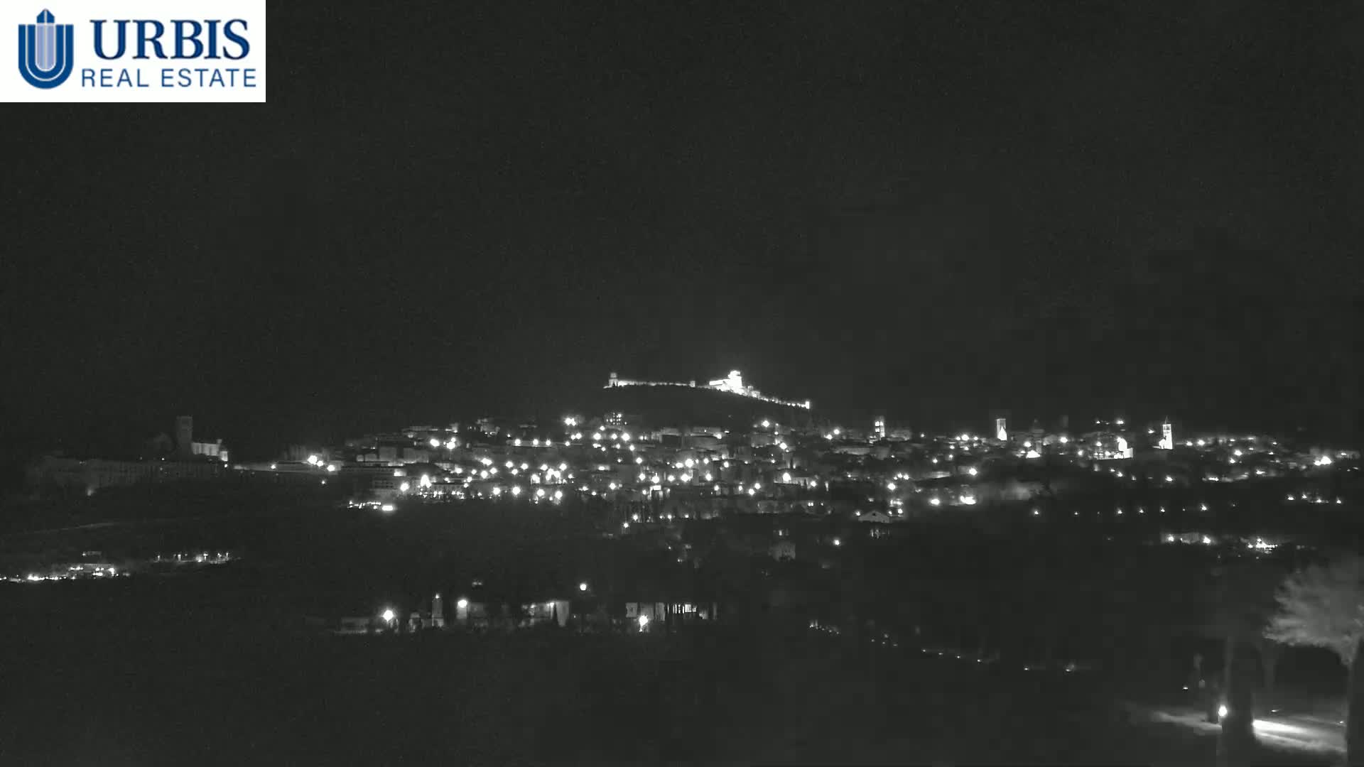 A nighttime view of a town illuminated by scattered lights, situated on a hill with a structure atop it under a clear, dark sky.
