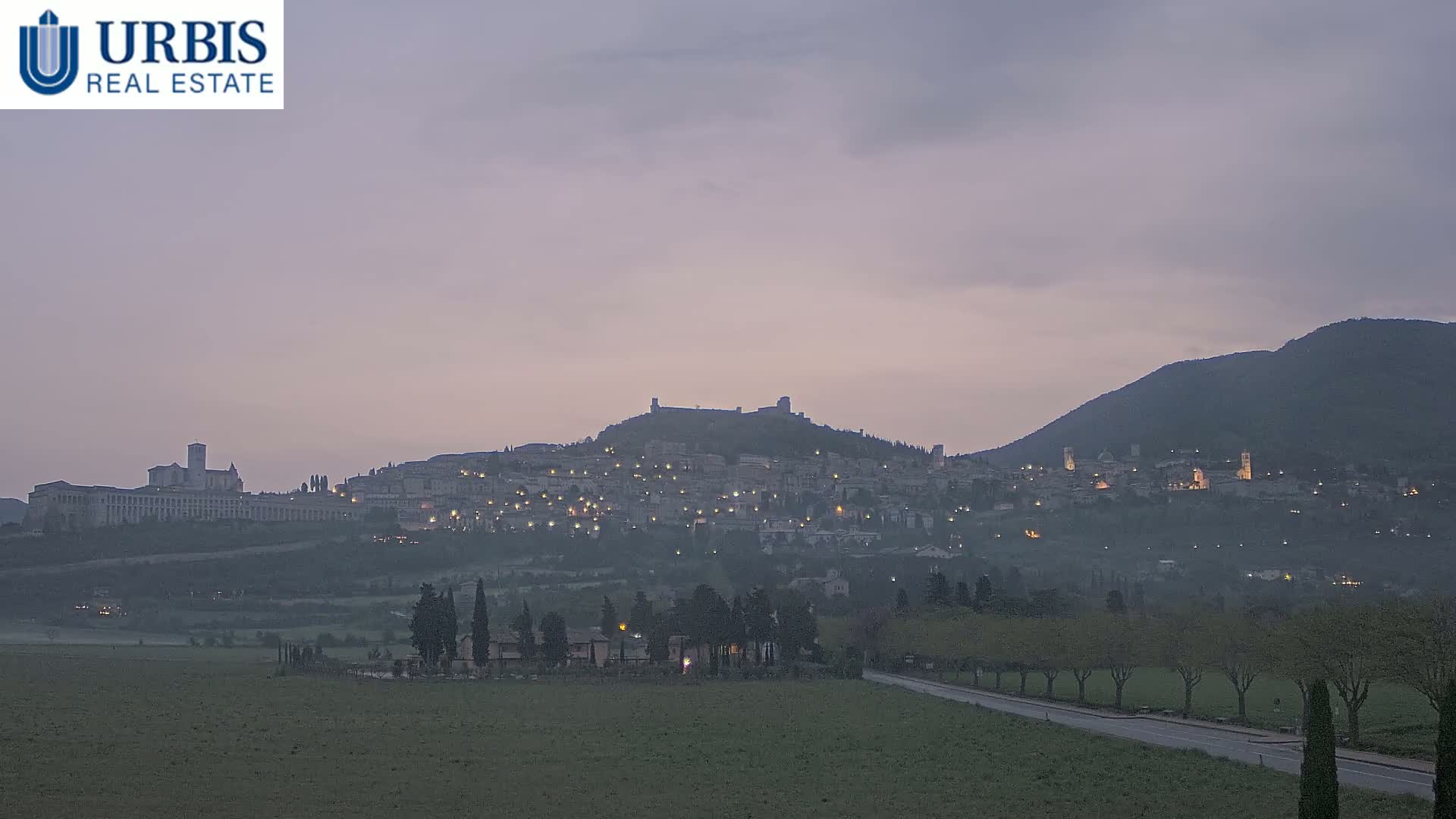 A hilltop town with lights illuminated at dusk under a pale, overcast sky, viewed from a distance across a flat field.