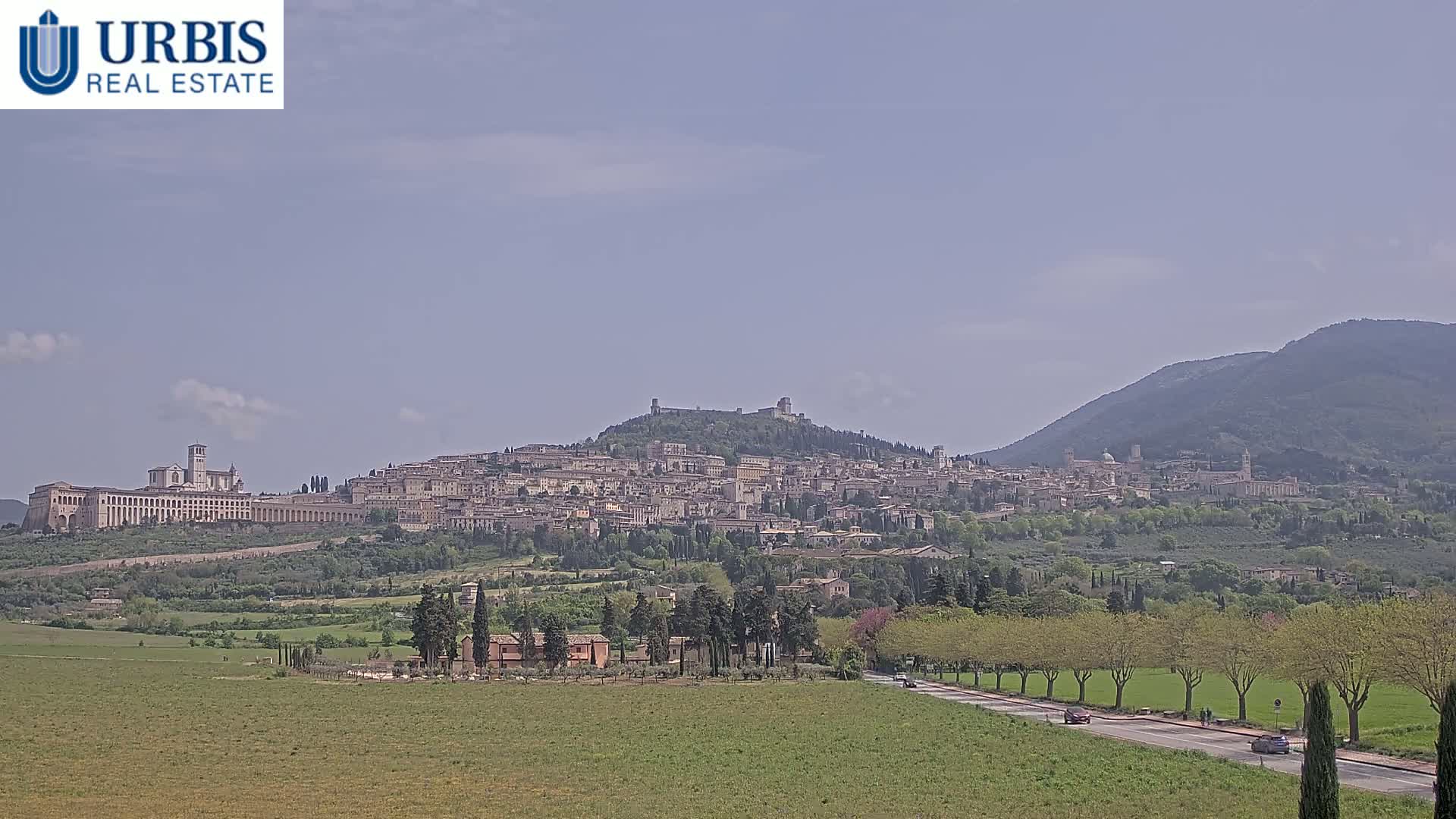 A hilltop town, nestled amongst green hills and under a mostly clear sky, is viewed from a distance across a flat field and road.