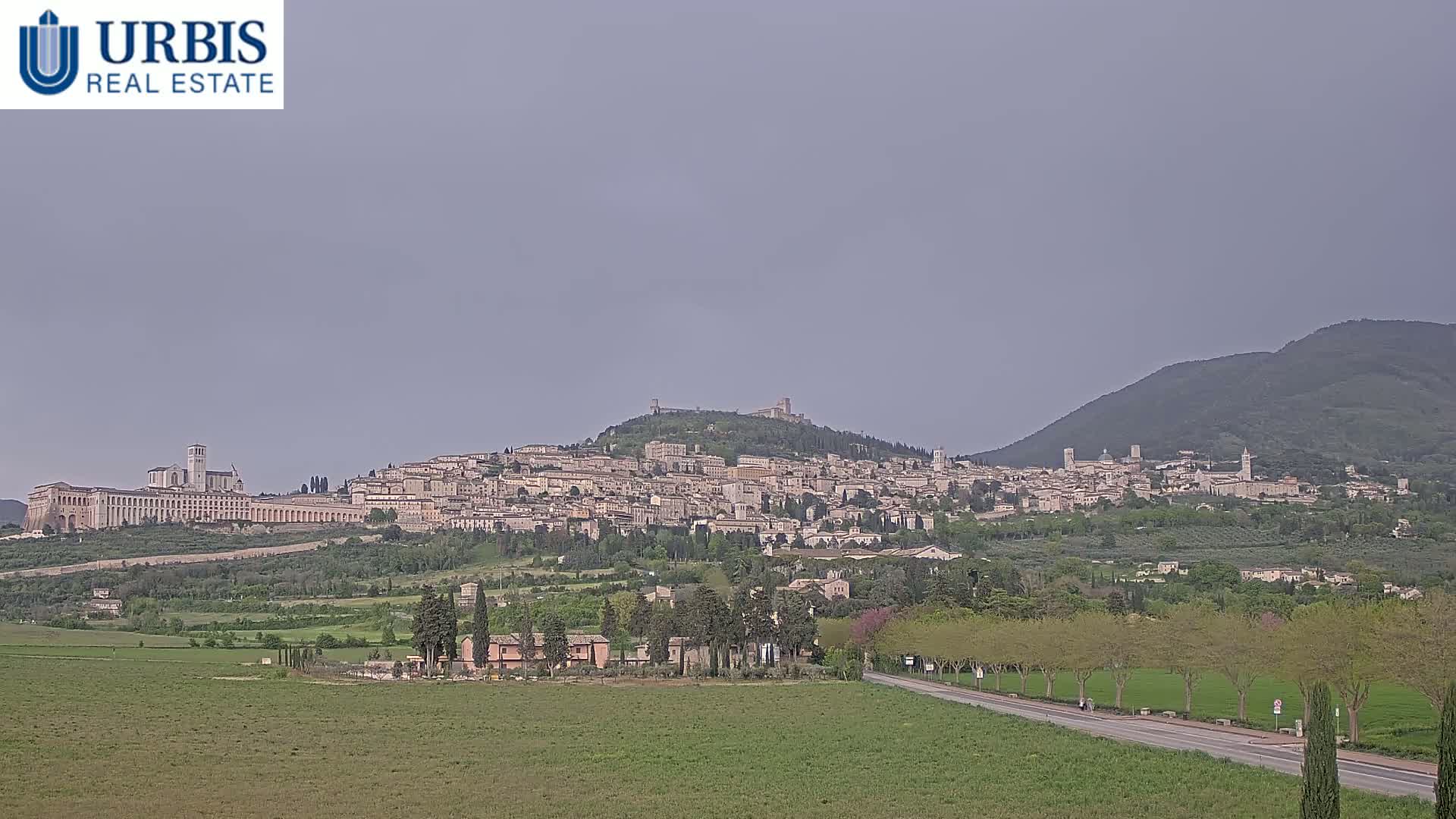 A hilltop town with numerous buildings, including a large basilica, is visible under an overcast sky, with green fields and trees in the foreground.