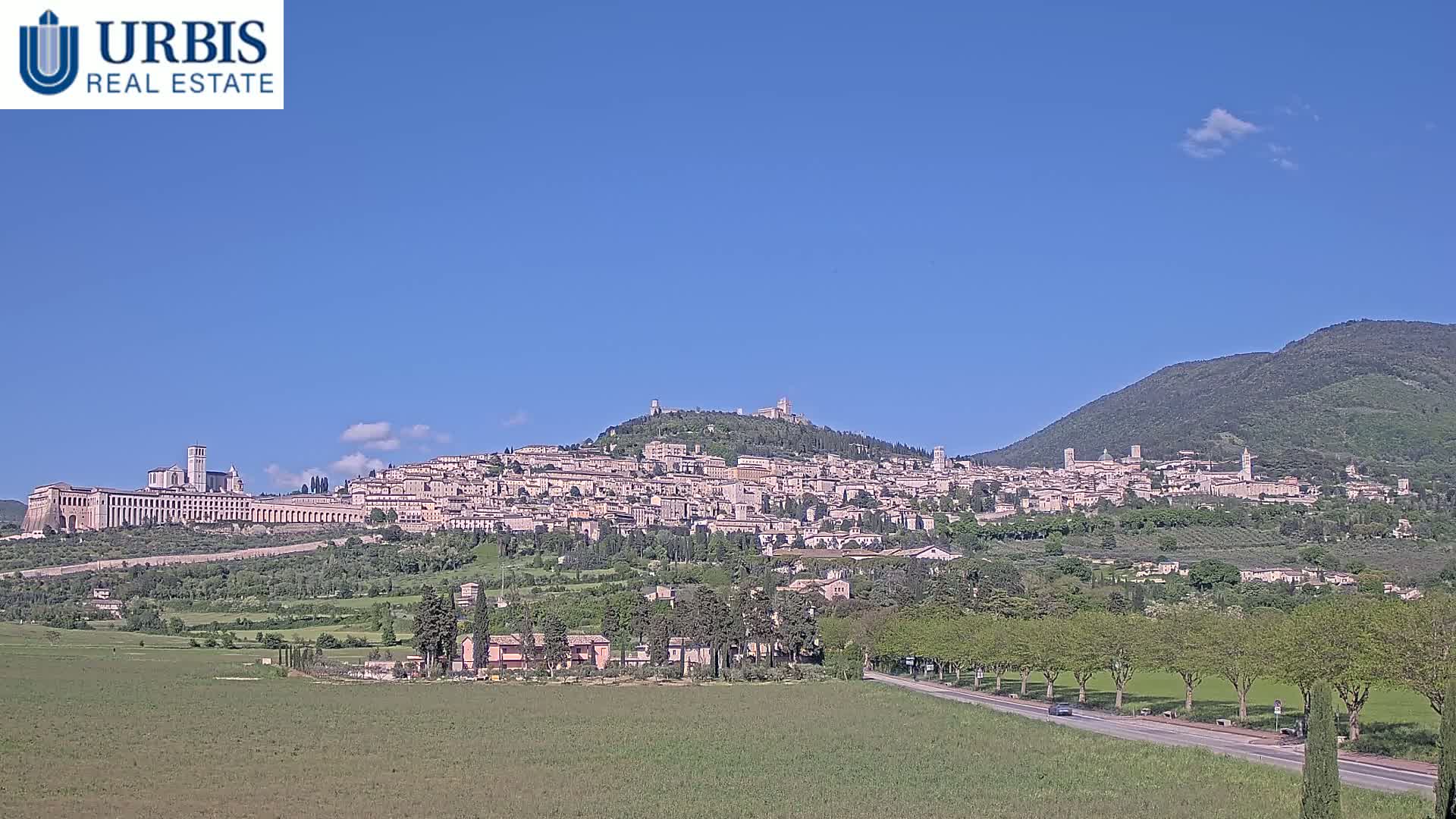A large hilltop town, under a clear blue sky with a few clouds, is surrounded by green fields and trees, with a road and a few cars visible in the foreground.