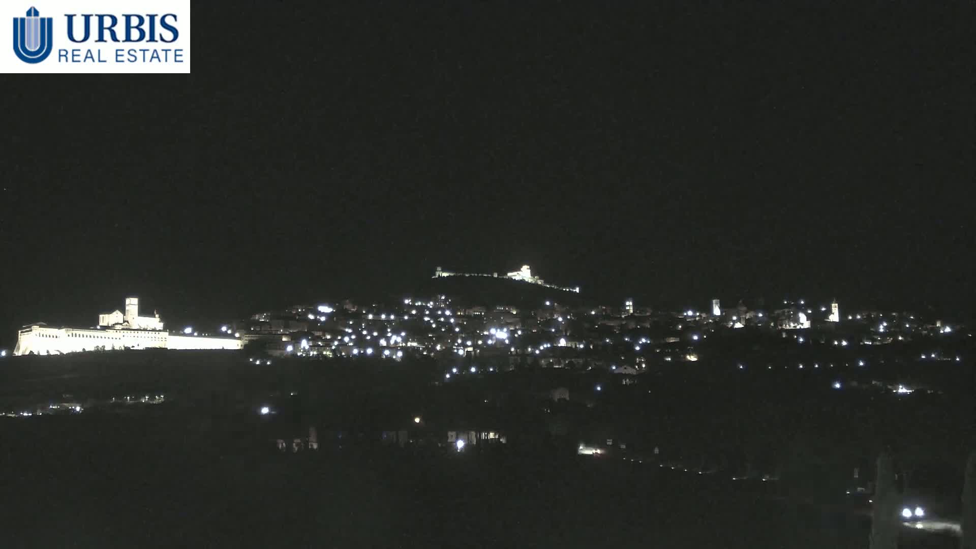 A nighttime view of a town nestled on a hill, illuminated by scattered lights, with a larger, brightly lit structure situated on a hilltop to the left.