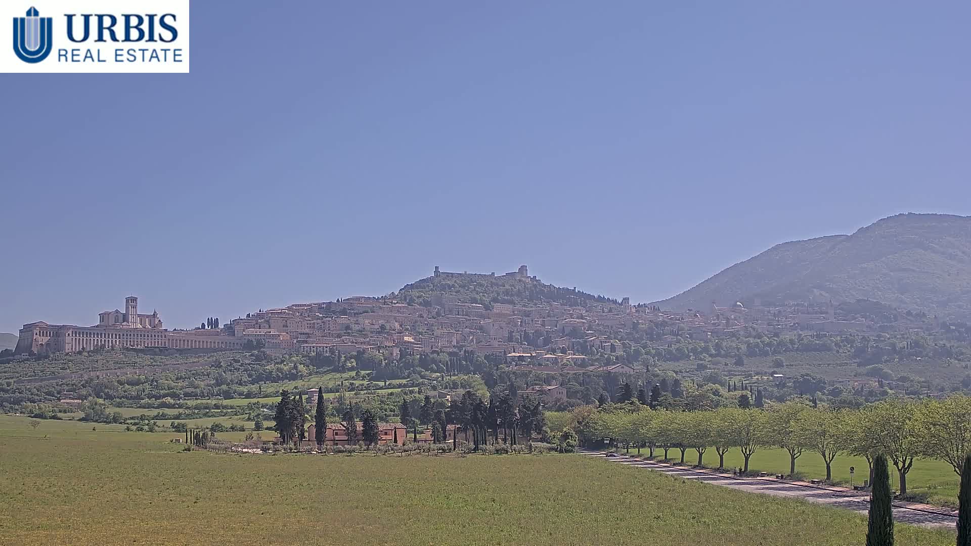 A hilltop town with a large basilica, nestled among rolling green hills under a clear blue sky.