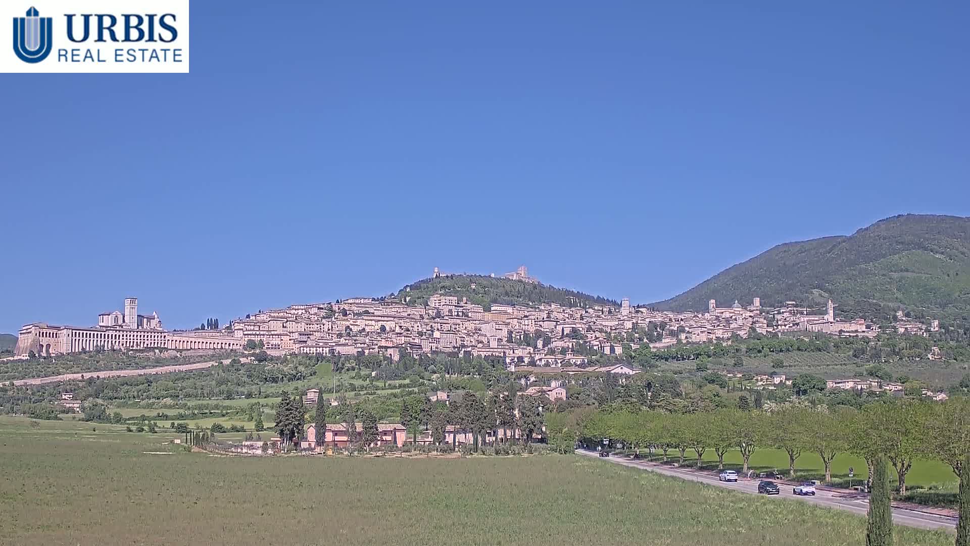 A large town sits atop a hill under a clear blue sky, with a long, flat building extending from its base, and cars driving on a road in the foreground.