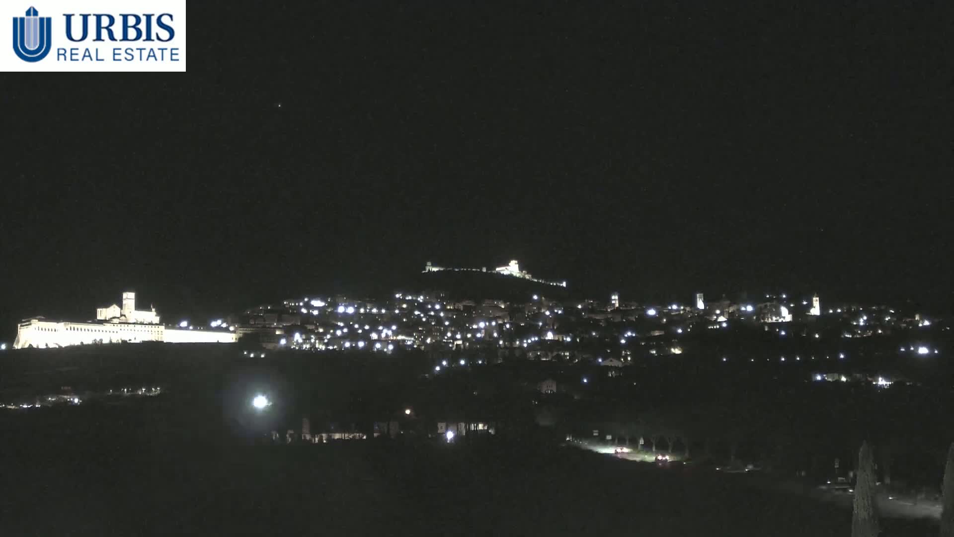 A nighttime view of a hilltop town illuminated by lights, with a large building complex visible on a lower hill to the left.