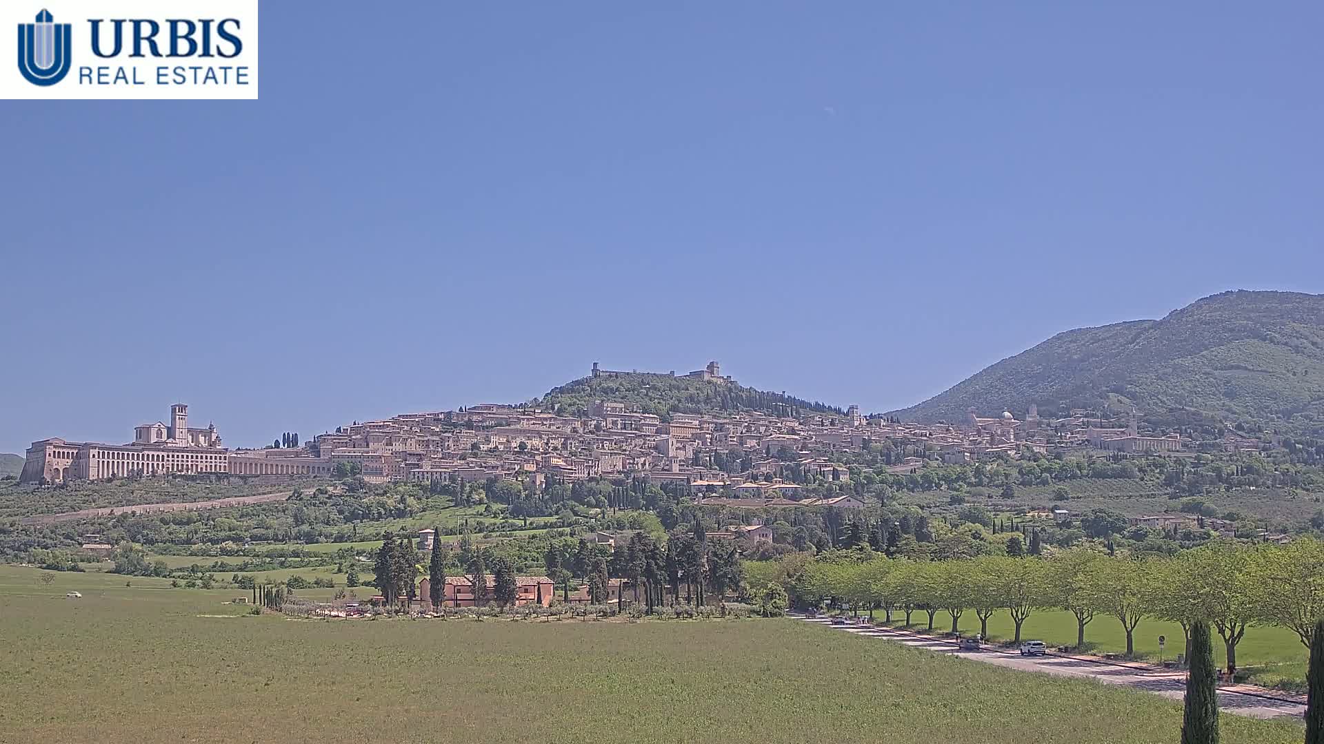 A large hill town, with a large basilica on its lower flank, is visible under a clear blue sky, and a road runs through a verdant field in the foreground.