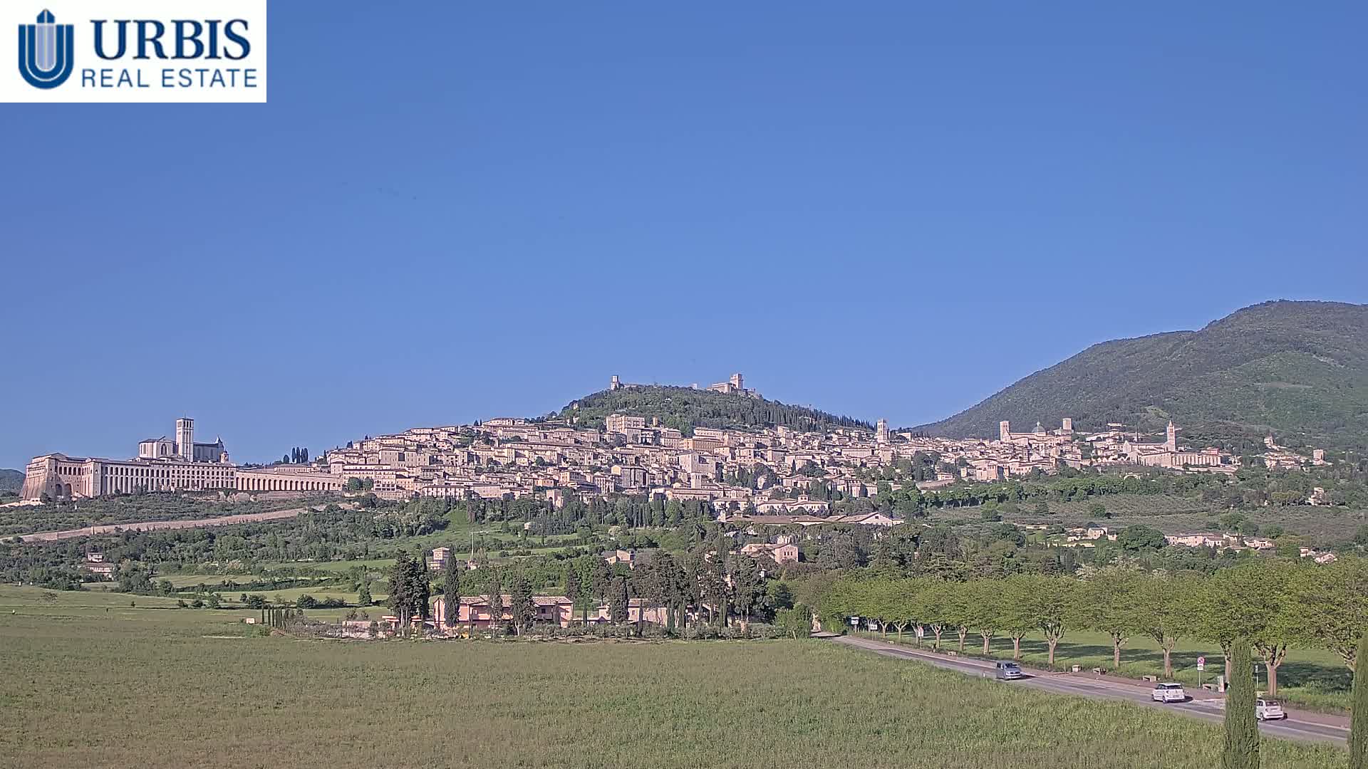 A large hill town is visible under a clear blue sky, with a road and green fields in the foreground.