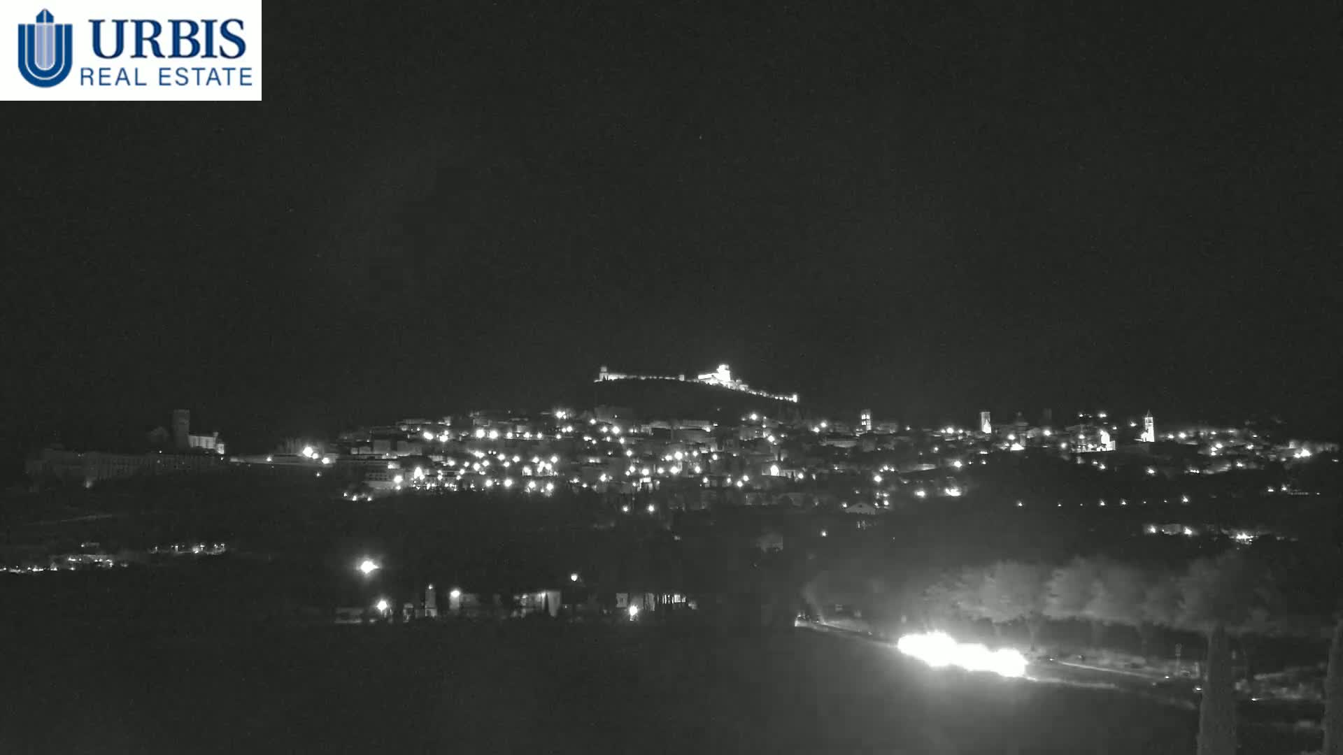 A nighttime long shot shows a hilltop town illuminated with scattered lights under a dark, clear sky.