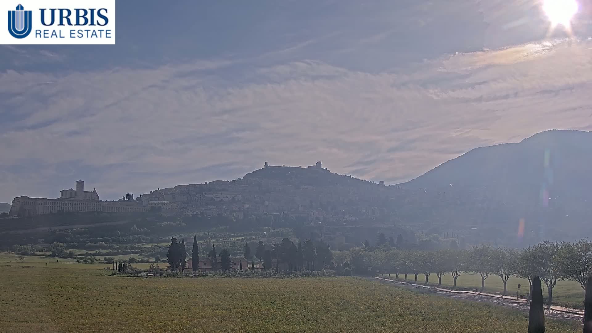 A hazy, sunlit view shows a hilltop town, including a large basilica, situated before a larger mountain range under a mostly cloudy sky.