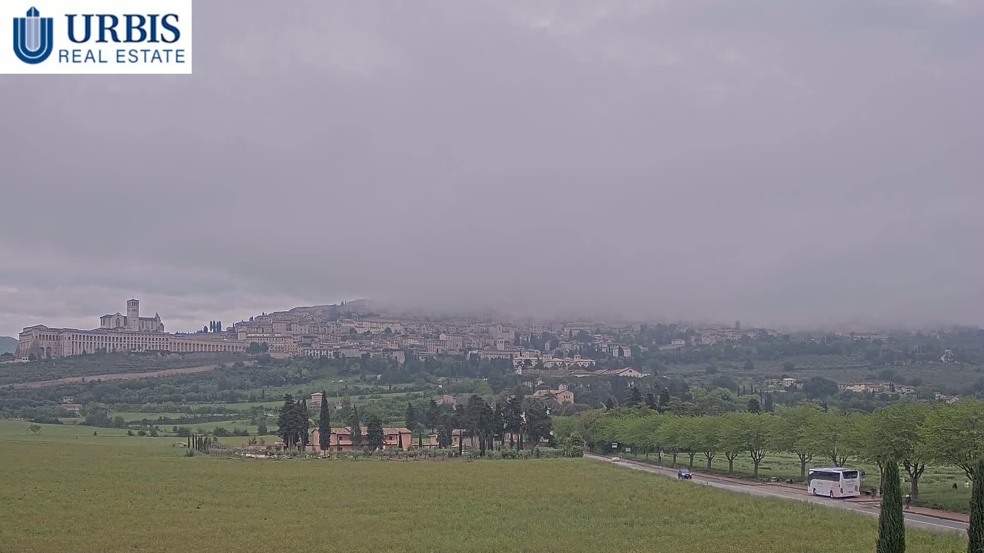 A large town sits atop a hill shrouded in fog, with a bus driving on a road in the foreground of a green field.