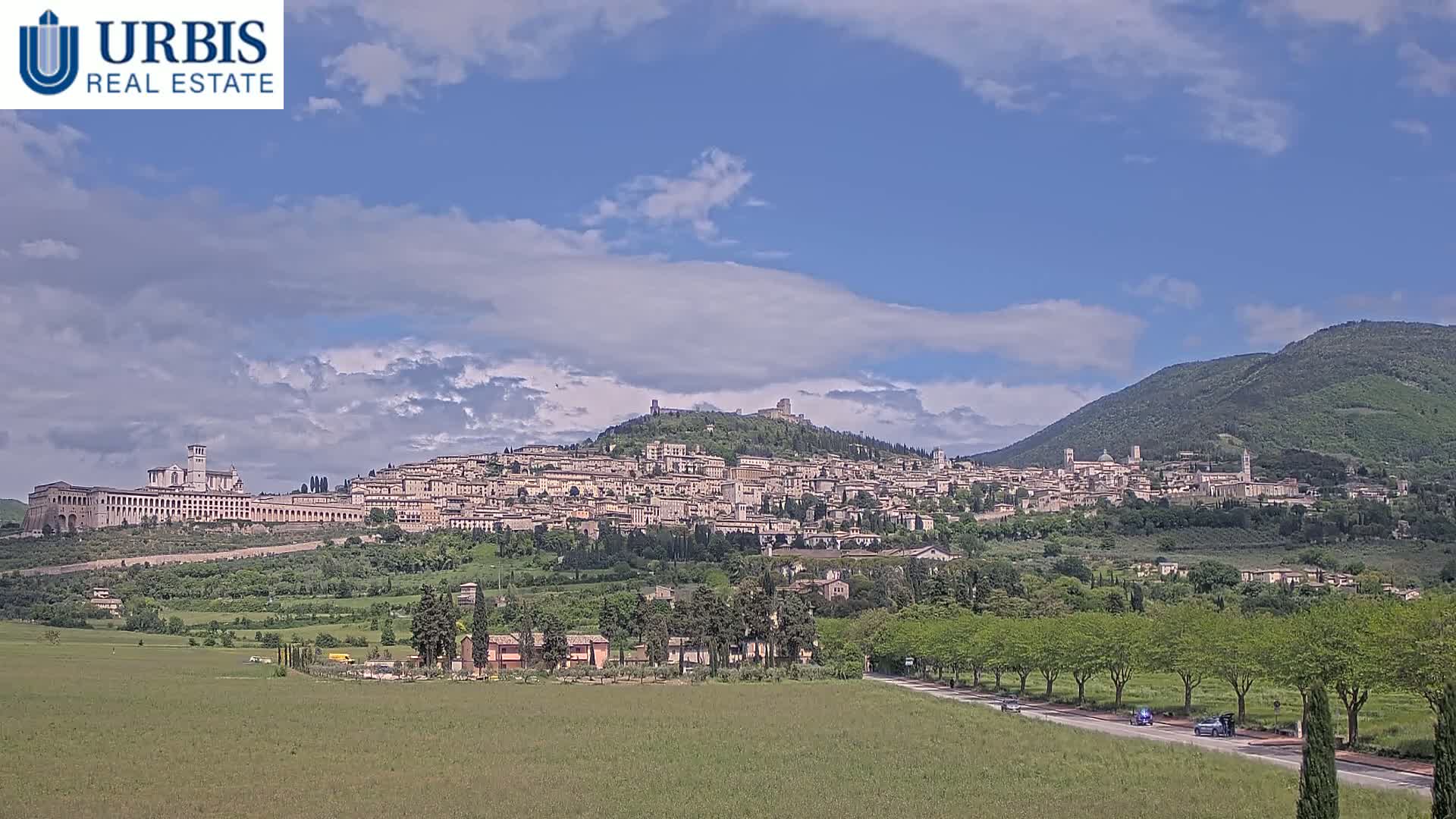 A large hilltop town, nestled between rolling green hills under a partly cloudy blue sky, is visible from a distance.