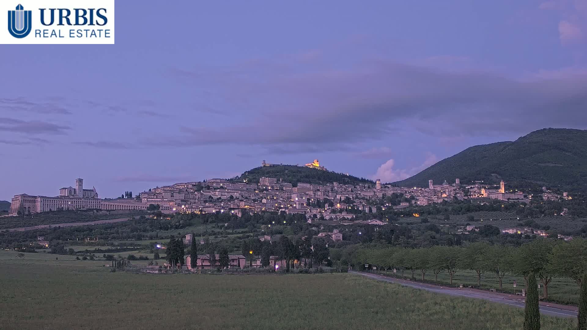 A hilltop town is illuminated at twilight under a mostly clear, dusky purple sky, with a large building complex at its base and a mountain in the background.