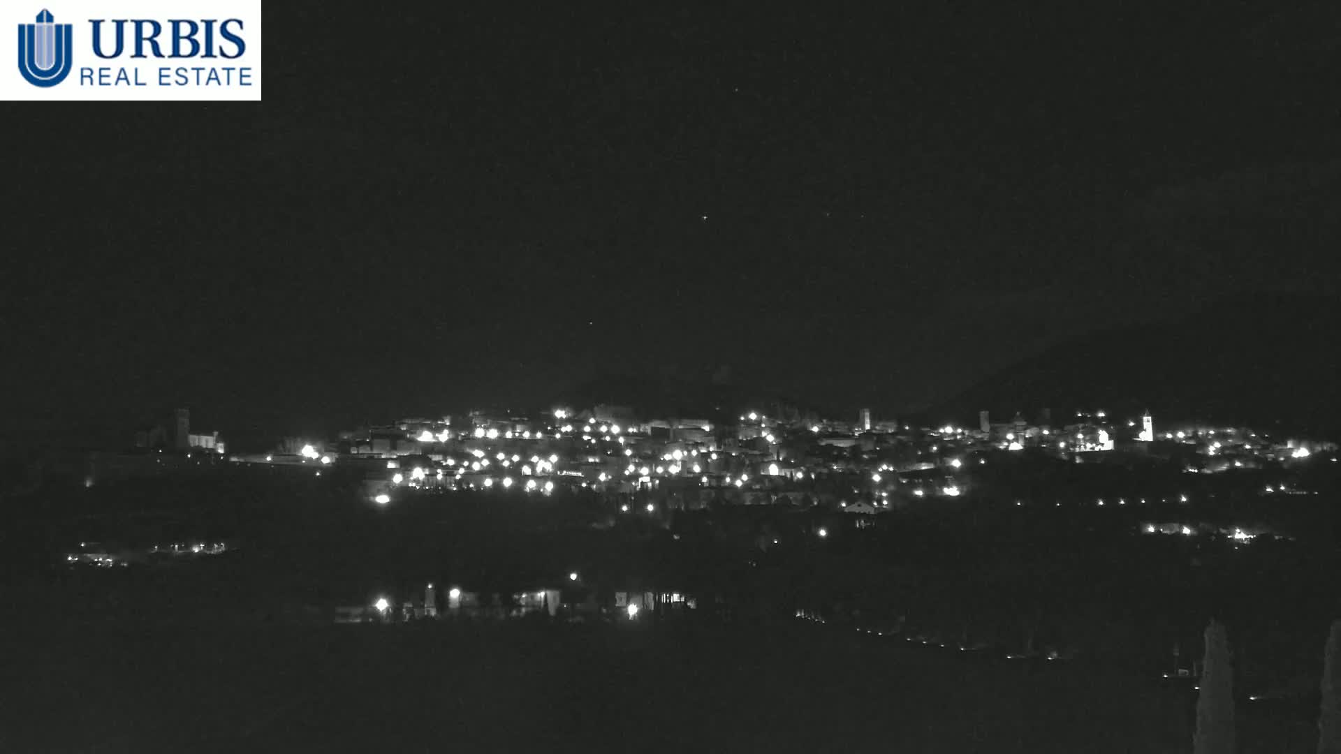 A nighttime, long-range view of a town nestled in a hillside, illuminated by scattered lights under a clear, dark sky.