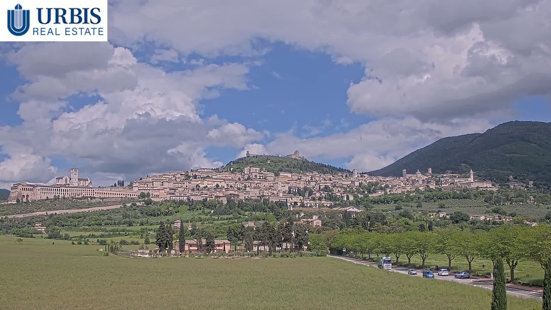 A hilltop town with a large basilica stretches across a verdant hillside under a partly cloudy sky.