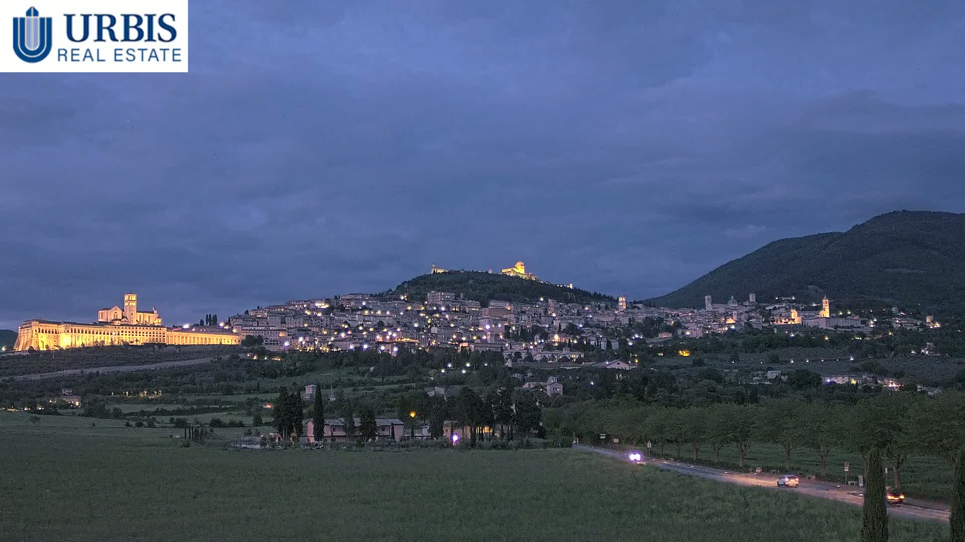A hilltop town illuminated at night under a twilight sky, with a large building extending along the base of the hill.