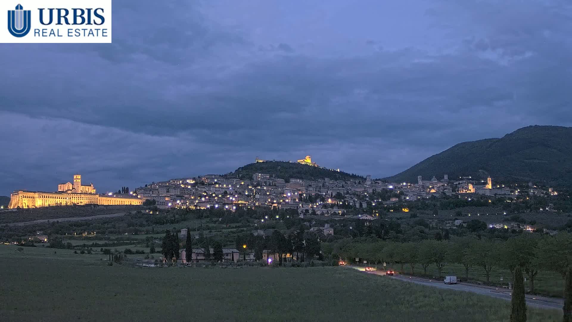 A hilltop town illuminated at night under a twilight sky, with a large building and a mountain in the background.