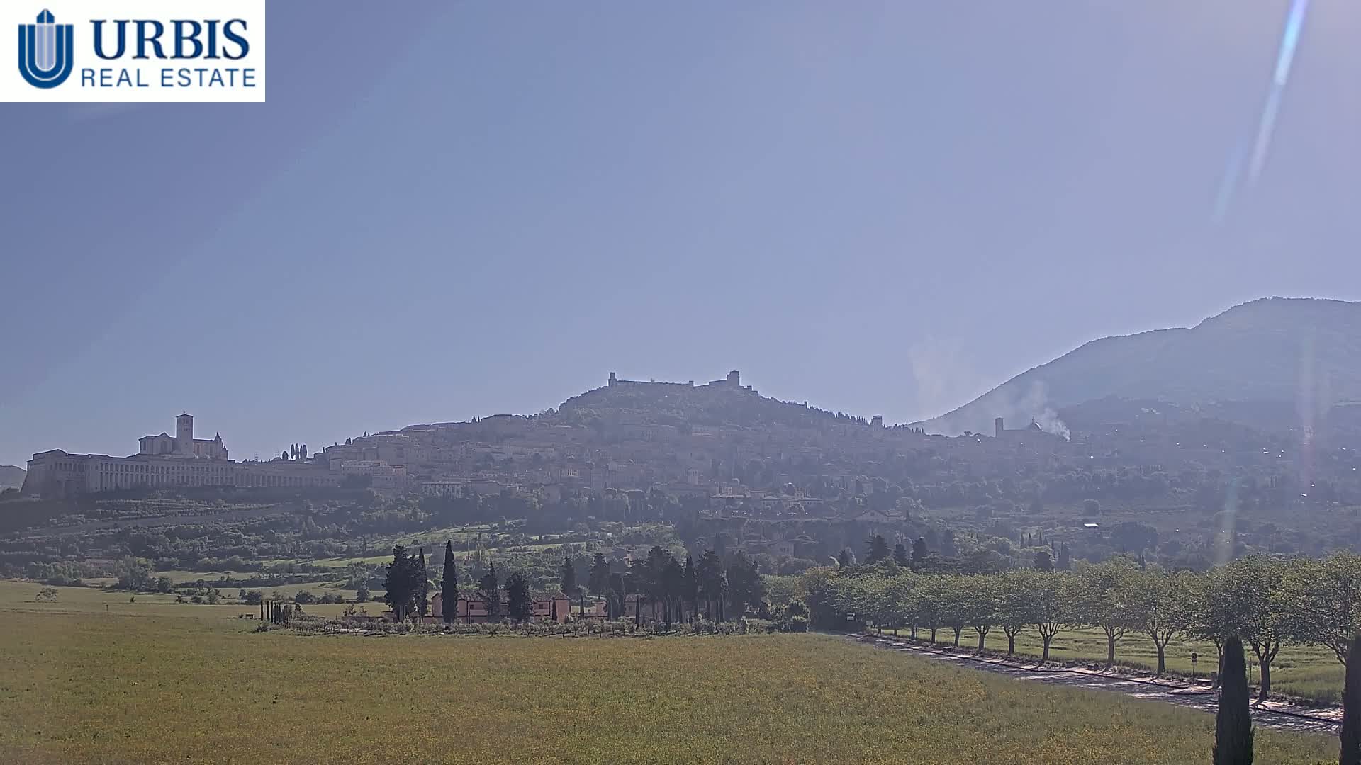 A hazy, distant view of a hilltop town, featuring a large church and fortress, stretches across a green field under a clear sky.