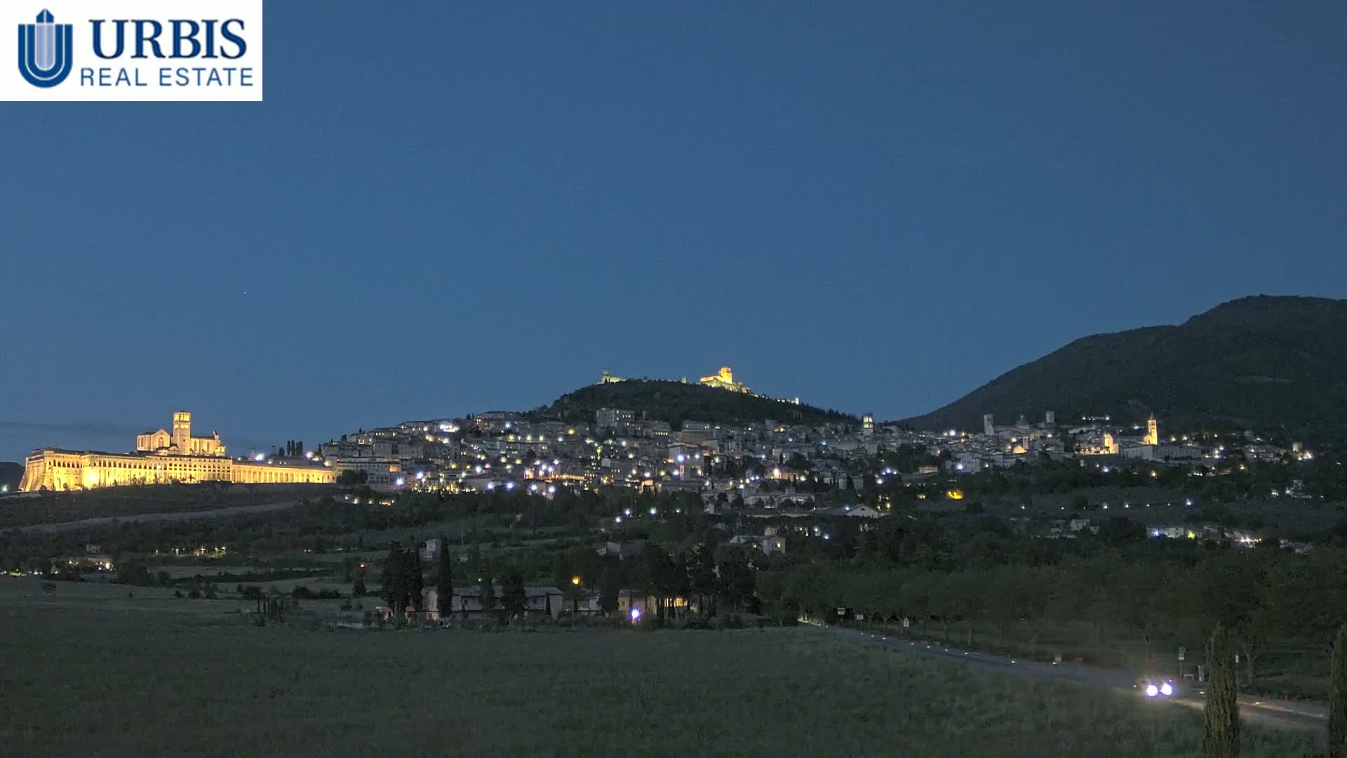 A hilltop town illuminated at night, with a large basilica visible in the foreground, under a clear, dark blue sky.