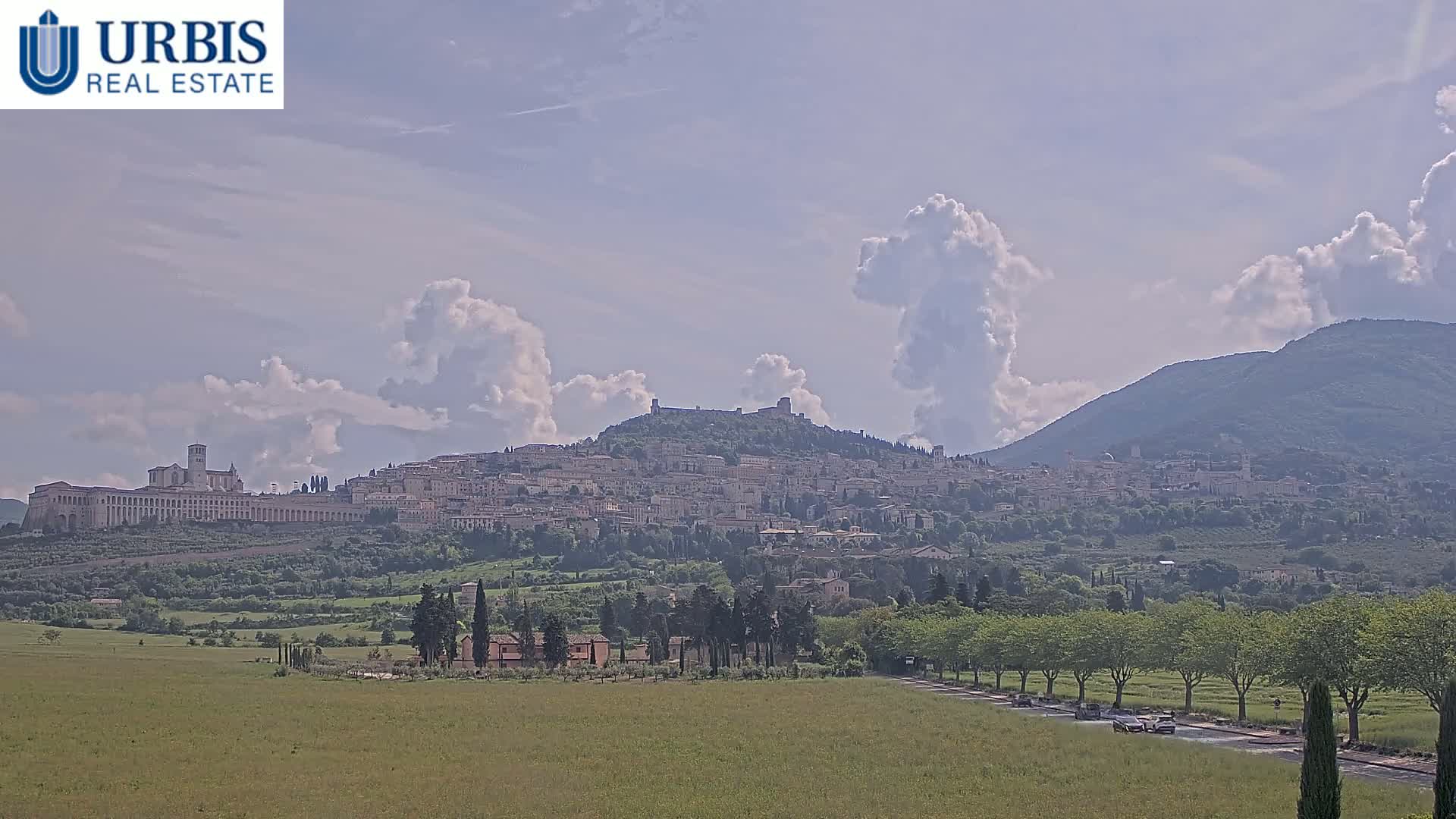 A hilltop town, partly obscured by partly cloudy skies, is visible in the distance, with a flat, green field in the foreground.