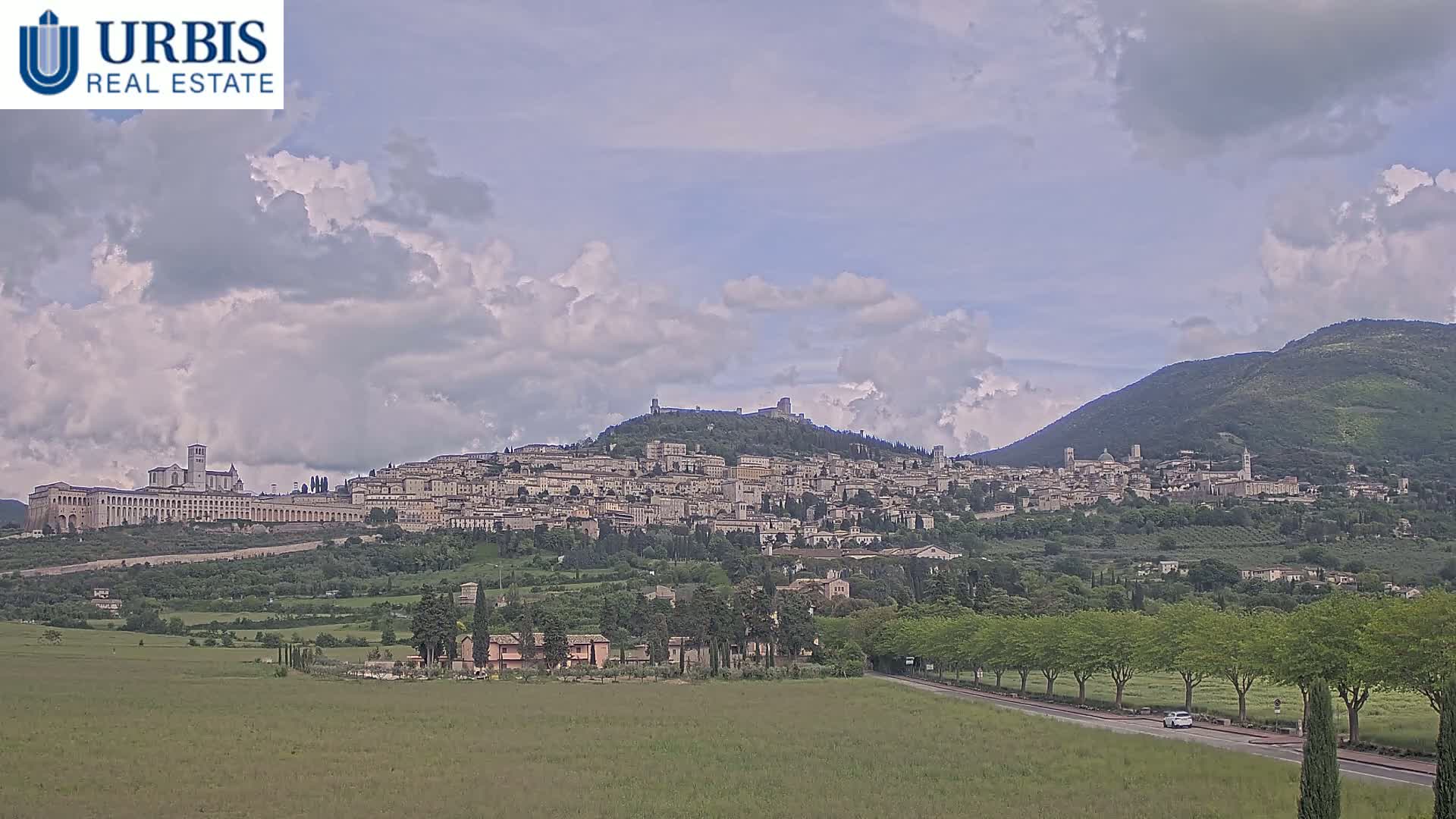 A sprawling hilltop town, nestled among green hills under a partly cloudy sky, is seen from a distance.
