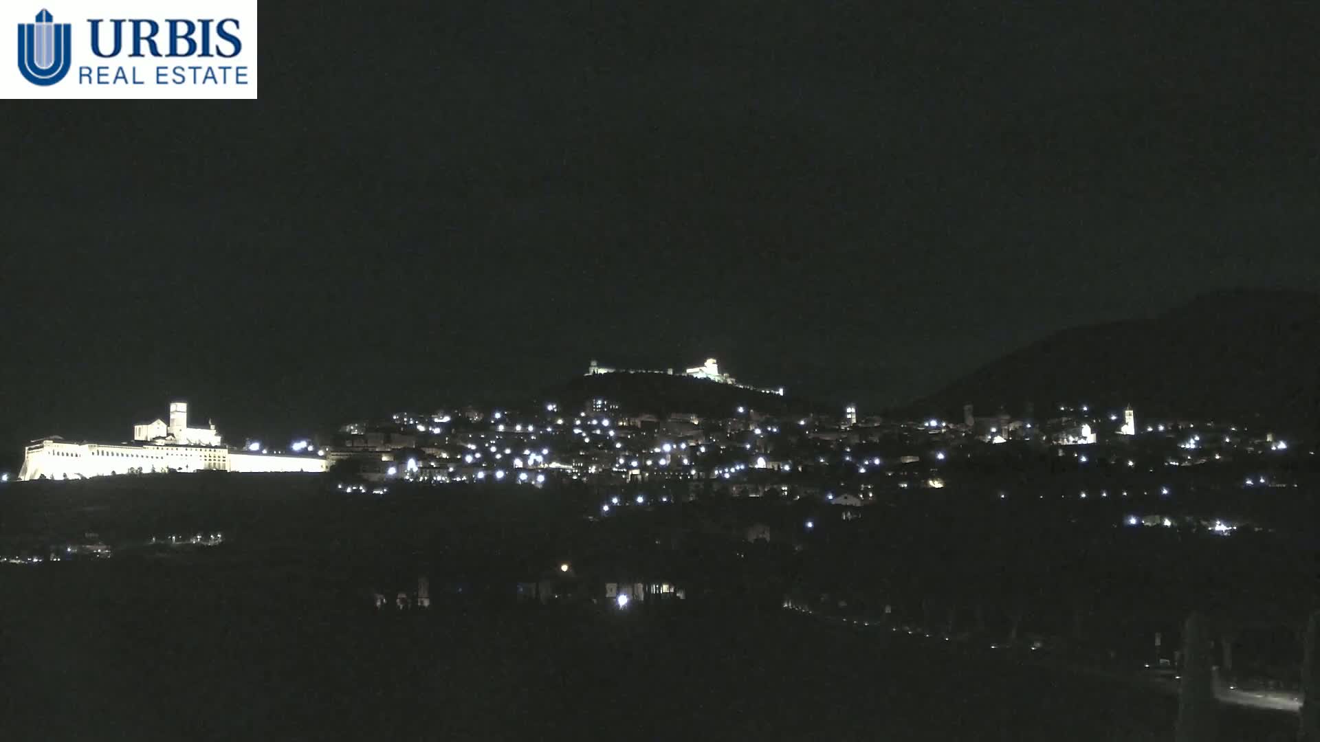 A nighttime view shows a hilltop town illuminated by scattered lights, with a large, brightly lit structure on a lower hill in the foreground.