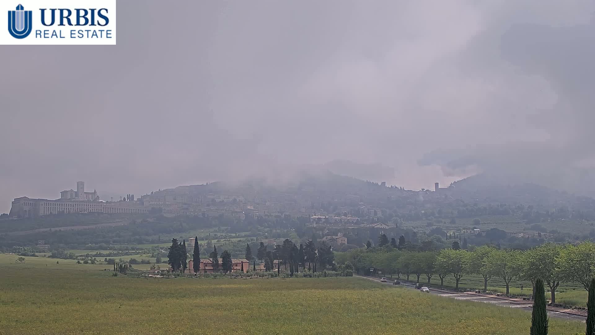 A hazy, distant view of a city nestled on a hill, with a large building prominent in the foreground,  overlooking a grassy plain with trees and a road.