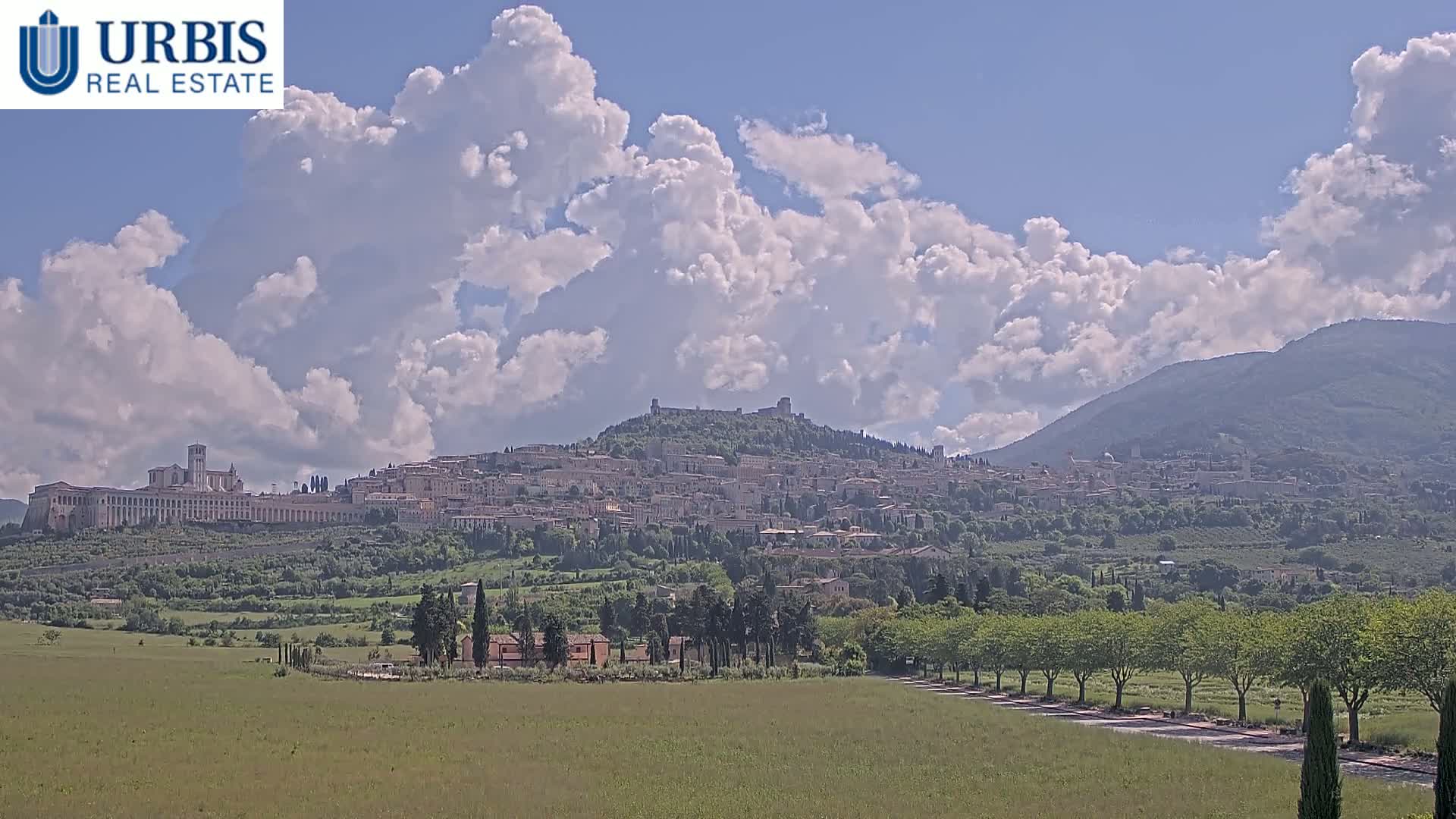 A hilltop town with a large basilica, nestled in a verdant valley under a partly cloudy sky, with mountains in the background.