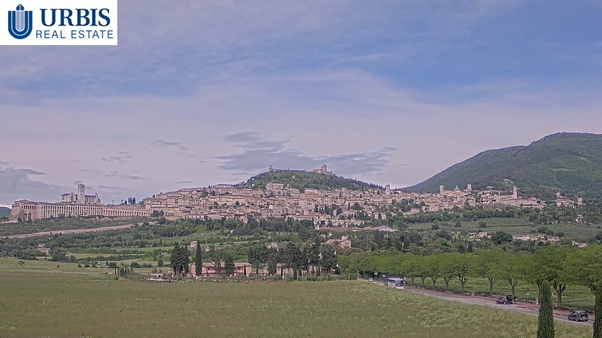 A large town is nestled on a hill, under a partly cloudy sky, with a green valley stretching into the foreground.