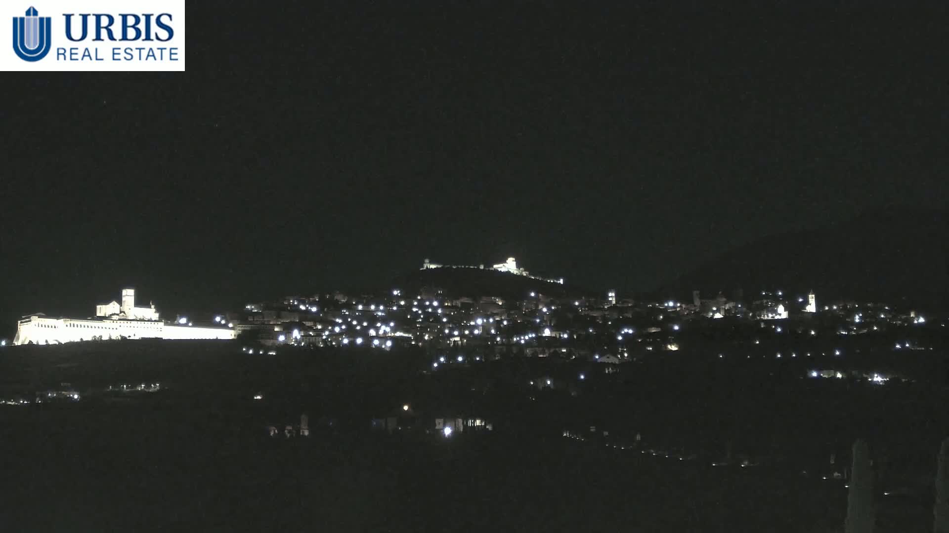 A nighttime long shot shows a hilltop town and a large, illuminated building complex at the base of a hill under a clear, dark sky.