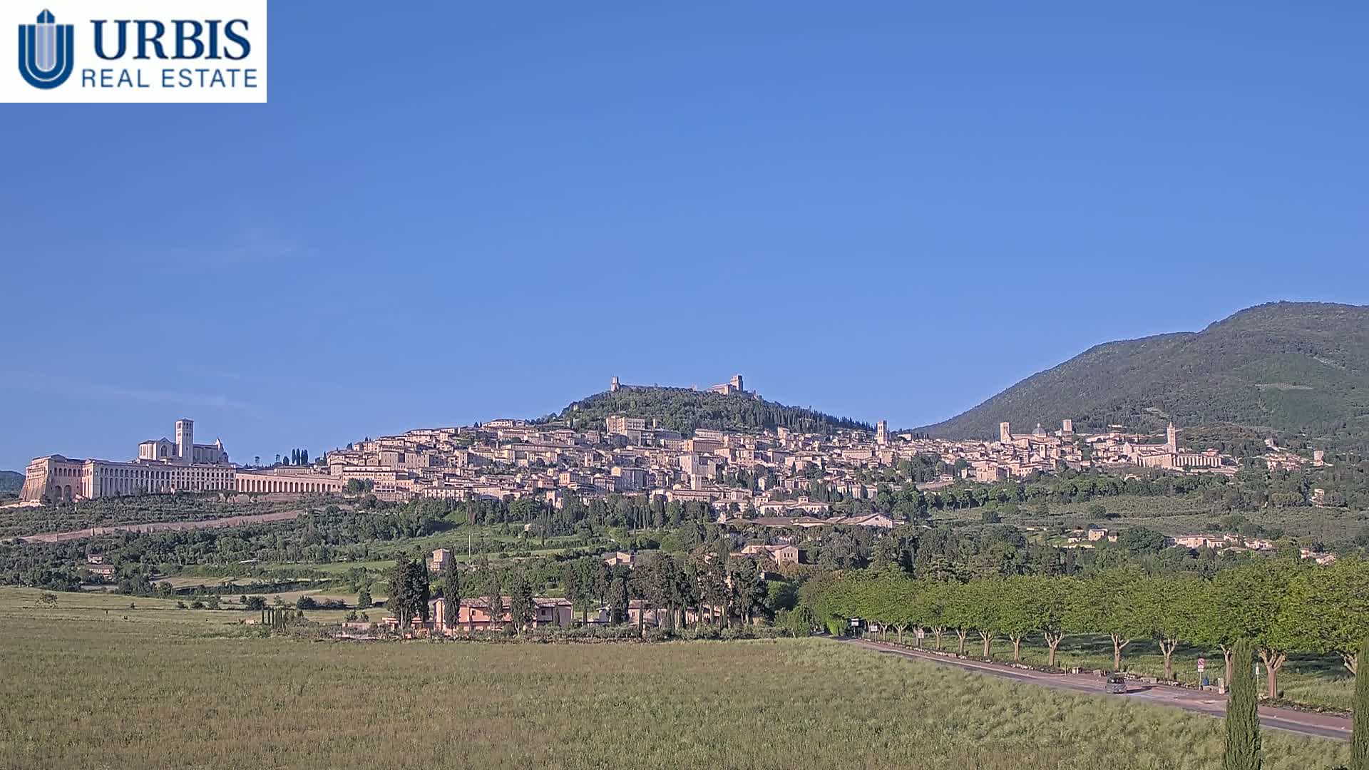 A hilltop town with numerous buildings, including a large basilica, is visible under a clear blue sky, with green fields and trees in the foreground.