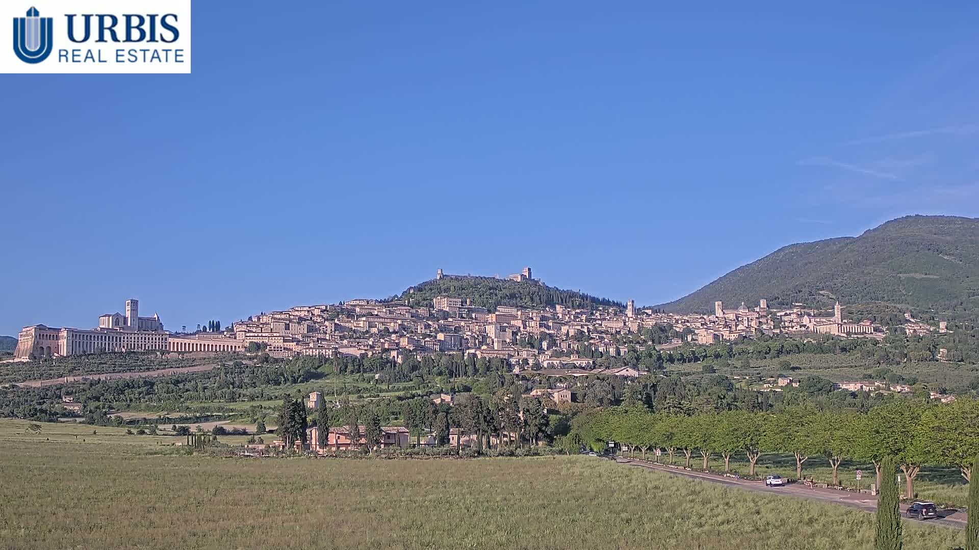 A hilltop town, nestled amongst green hills under a clear blue sky, is viewed from a distance across a flat field.