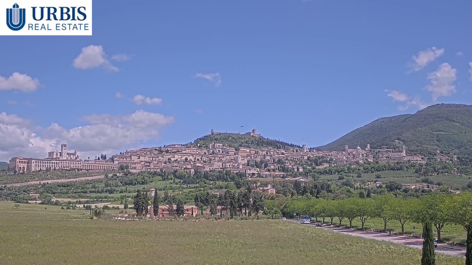 A hilltop town, nestled among green hills under a mostly sunny sky, stretches across a wide landscape.