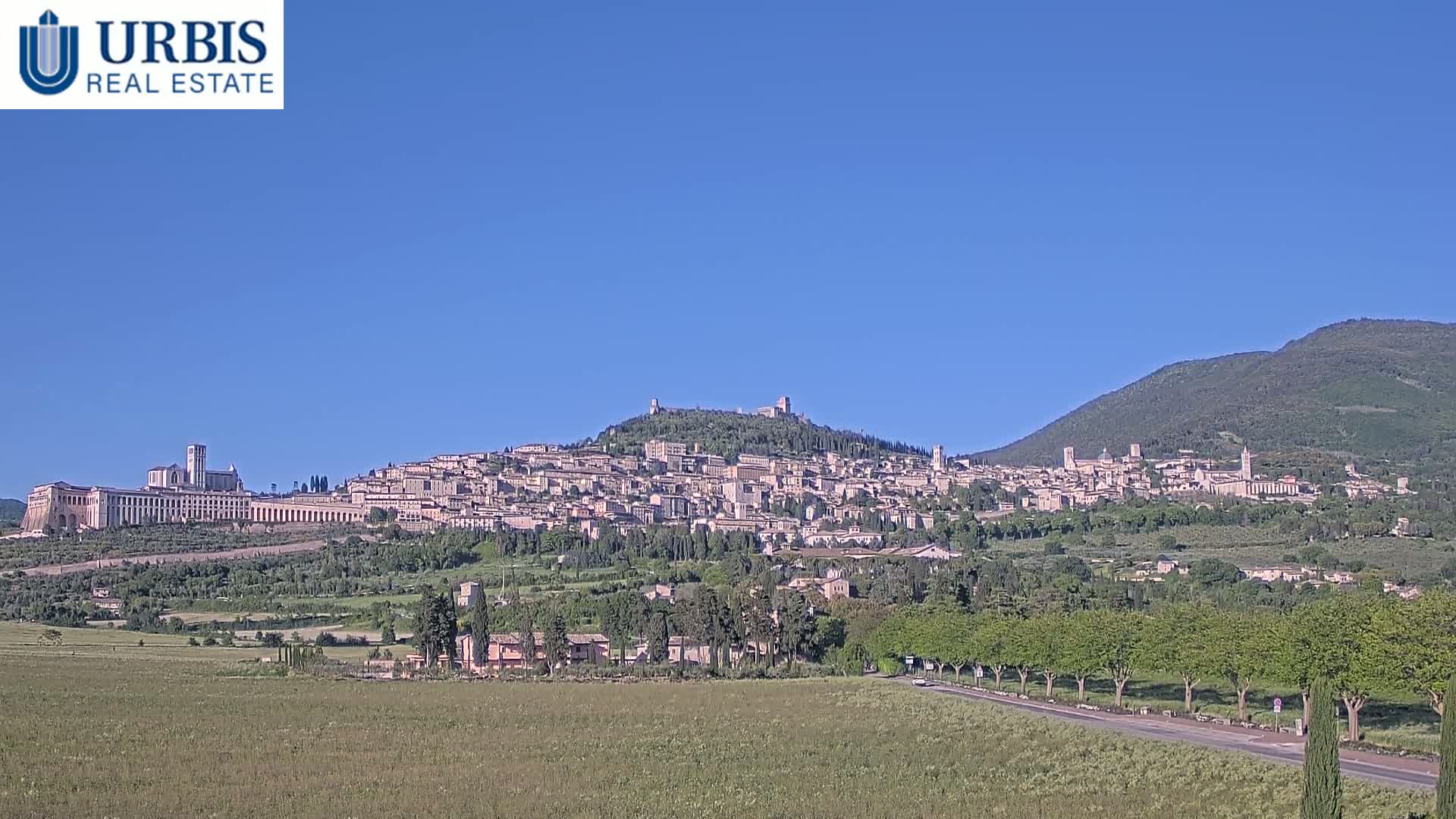 A hilltop town, nestled between green fields and mountains, is viewed under a clear blue sky.