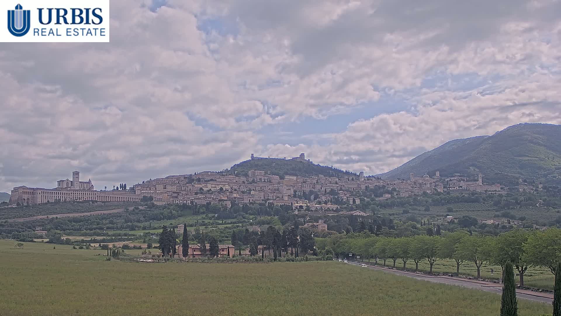 A hilltop town, nestled between mountains, is viewed from a distance under a partly cloudy sky.