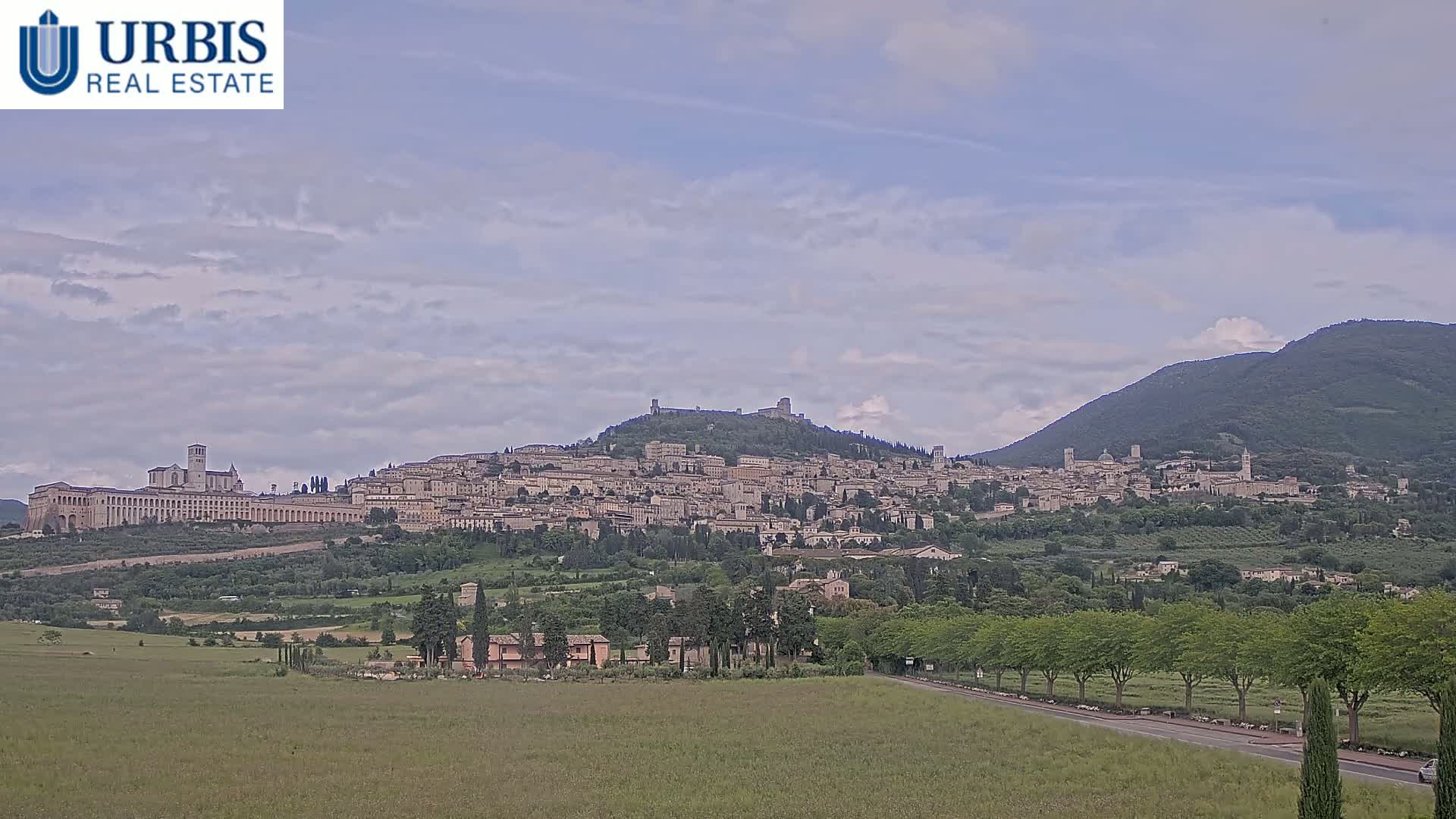 A hilltop town with numerous buildings, including a large basilica, is visible under a partly cloudy sky, with lush green vegetation in the foreground and a mountain in the background.