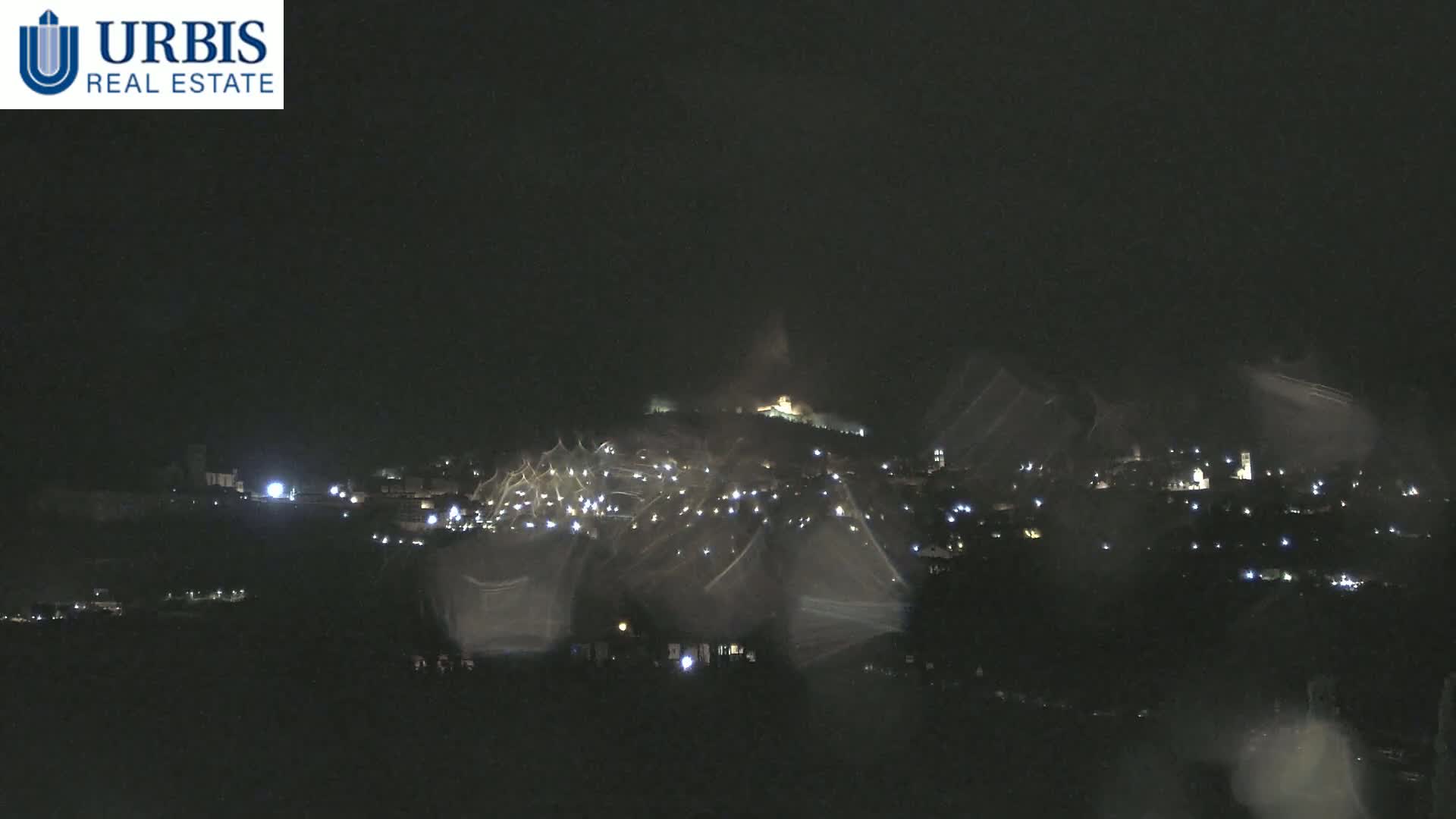 A nighttime view of a hilltop town with numerous lights visible through rain-streaked glass.
