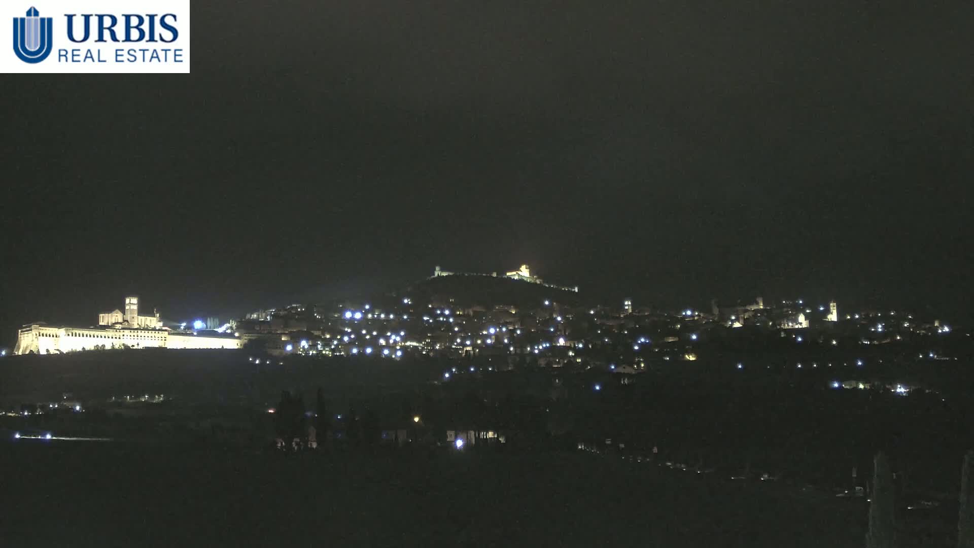 A nighttime long shot shows a hilltop town illuminated by many small lights, with a large, brightly lit building complex prominently featured at the bottom of the hill.