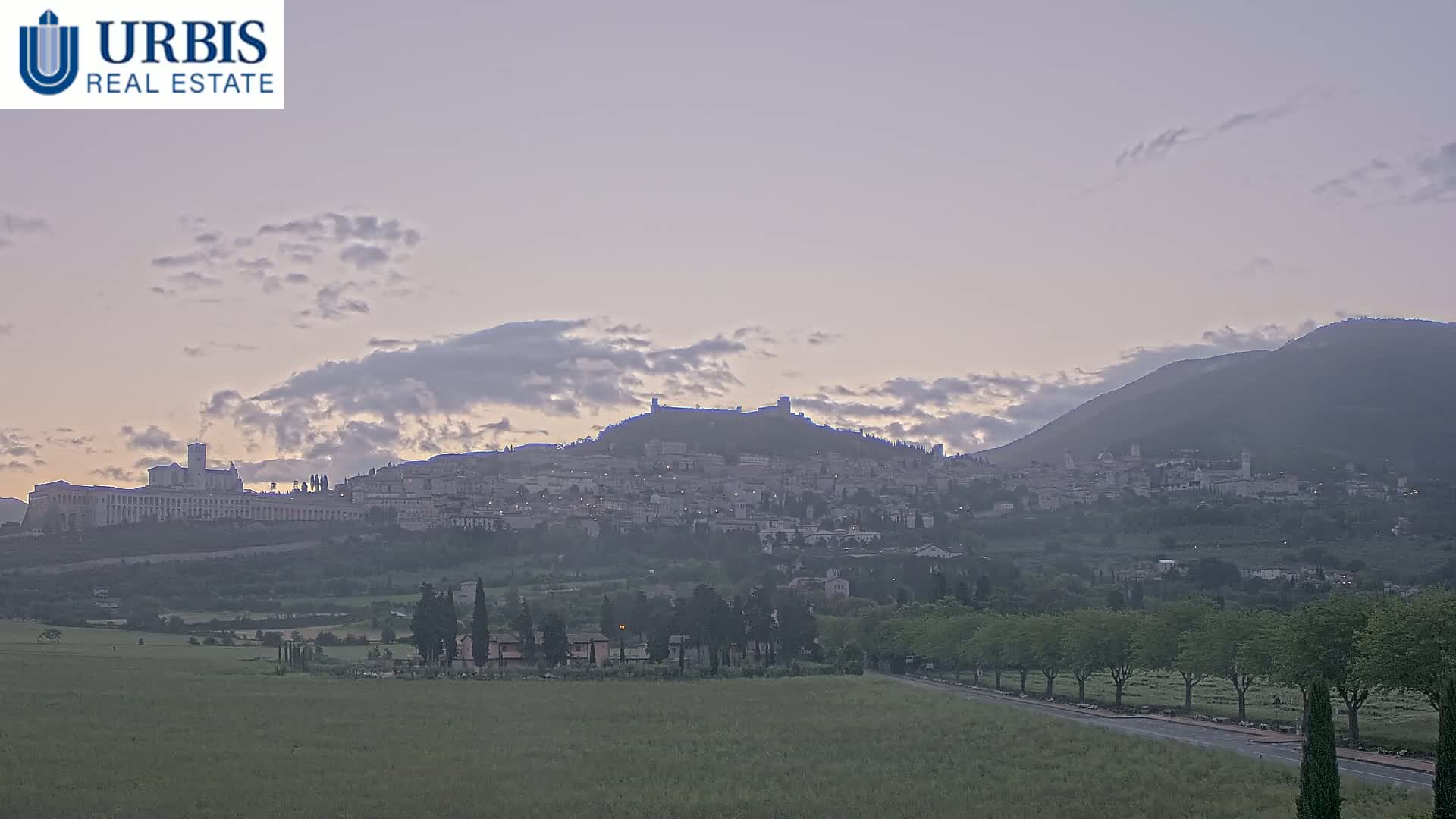 A hilltop town, possibly Assisi, is visible at dawn under a partly cloudy sky, with a large, flat plain in the foreground.