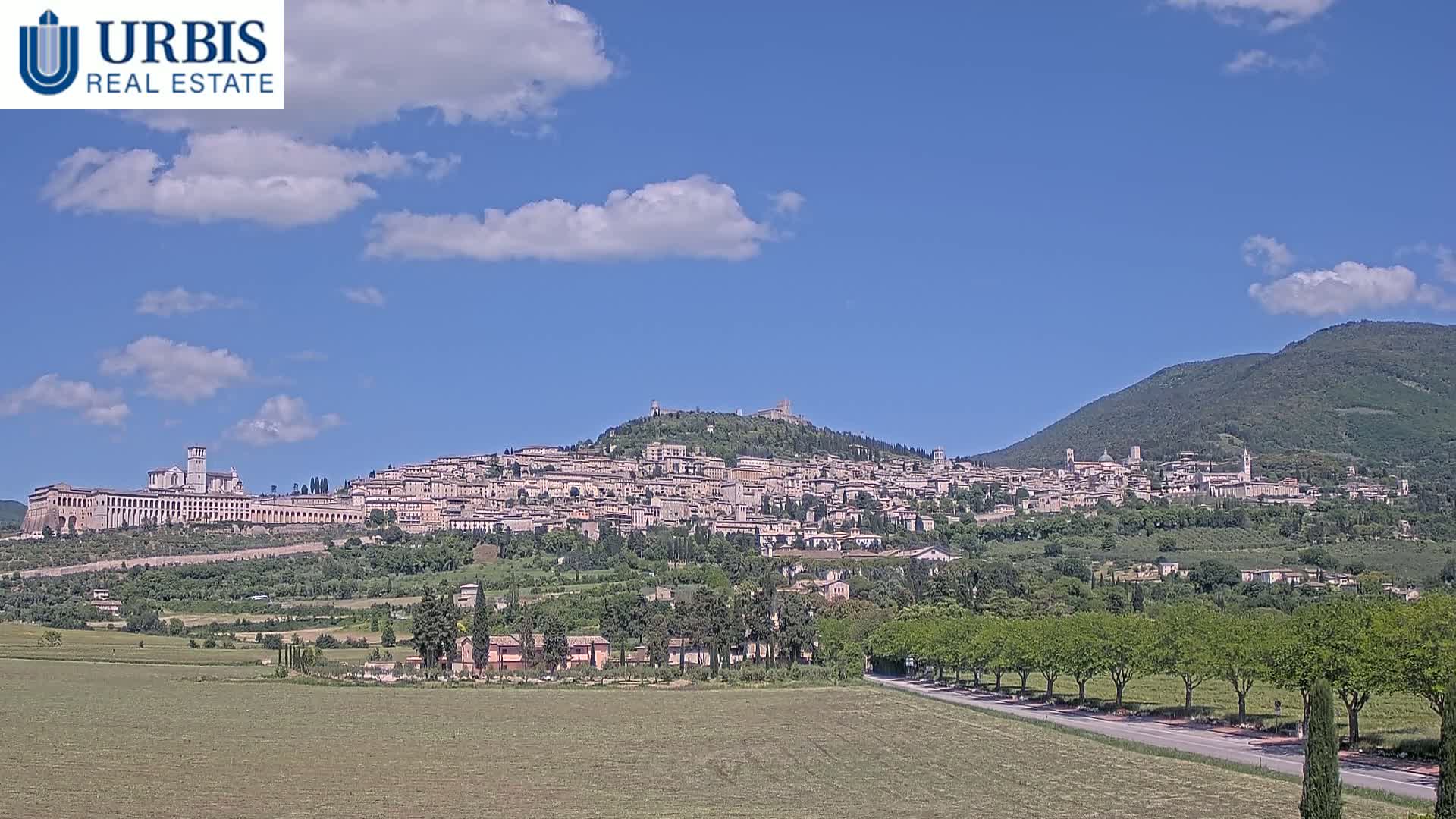 A hilltop town with numerous buildings, a large basilica, and surrounding green landscape is visible under a mostly sunny sky.
