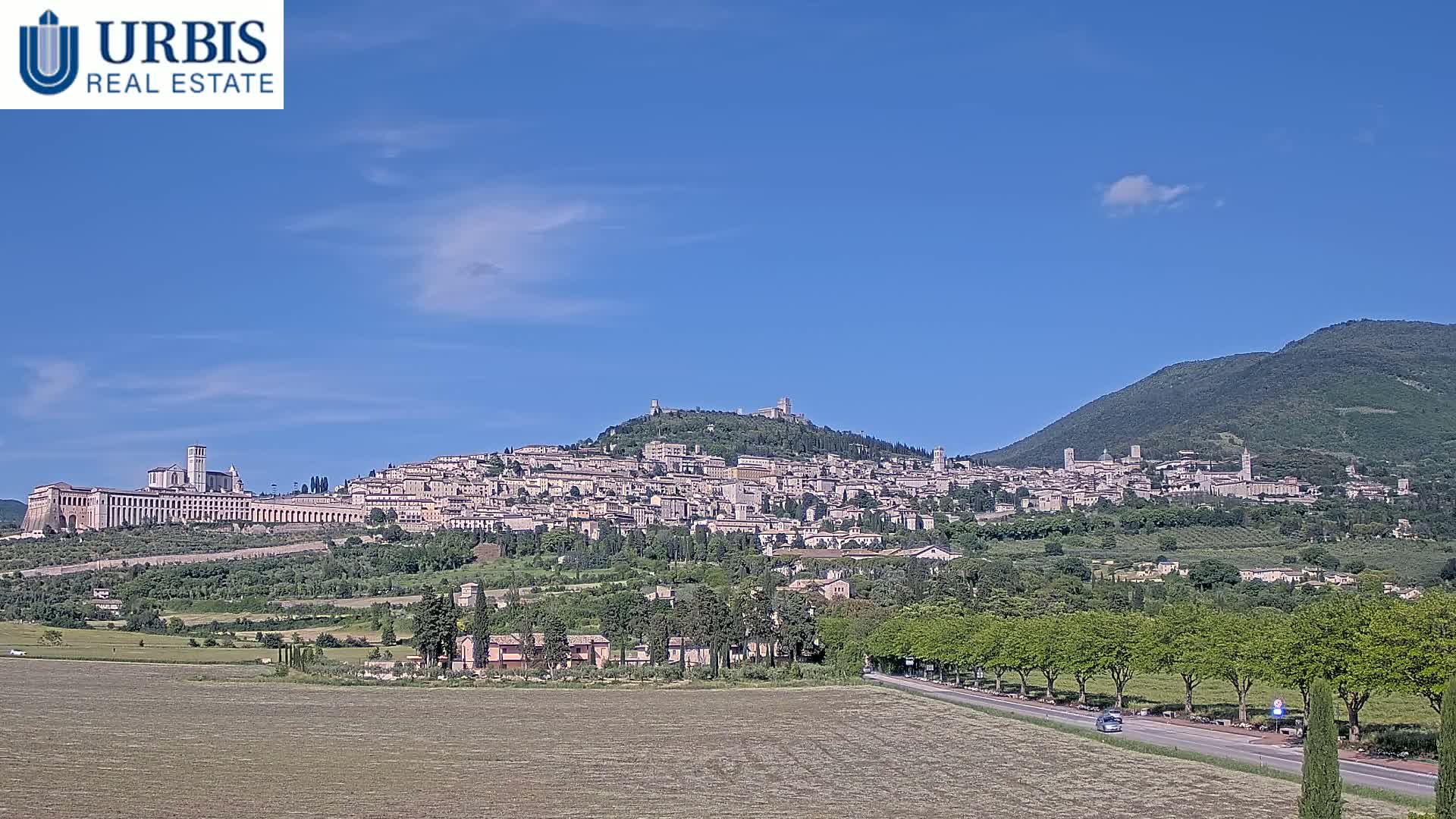 A sprawling hilltop town, under a clear blue sky, is backed by a larger mountain.