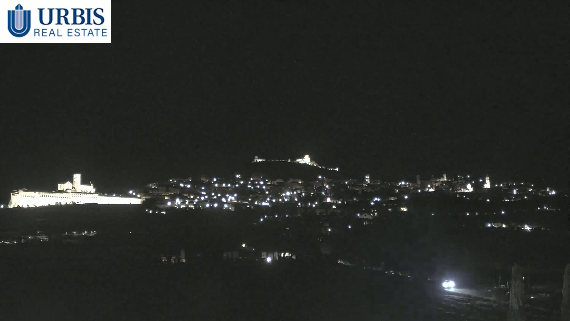 A nighttime view of a hilltop town illuminated by scattered lights, with a large, brightly lit structure visible on a lower hill in the distance.