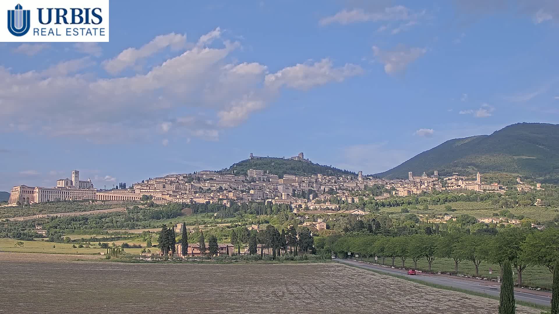 A sprawling hilltop town, possibly Assisi, under a mostly clear blue sky with scattered clouds, is seen from a distance across a verdant valley.