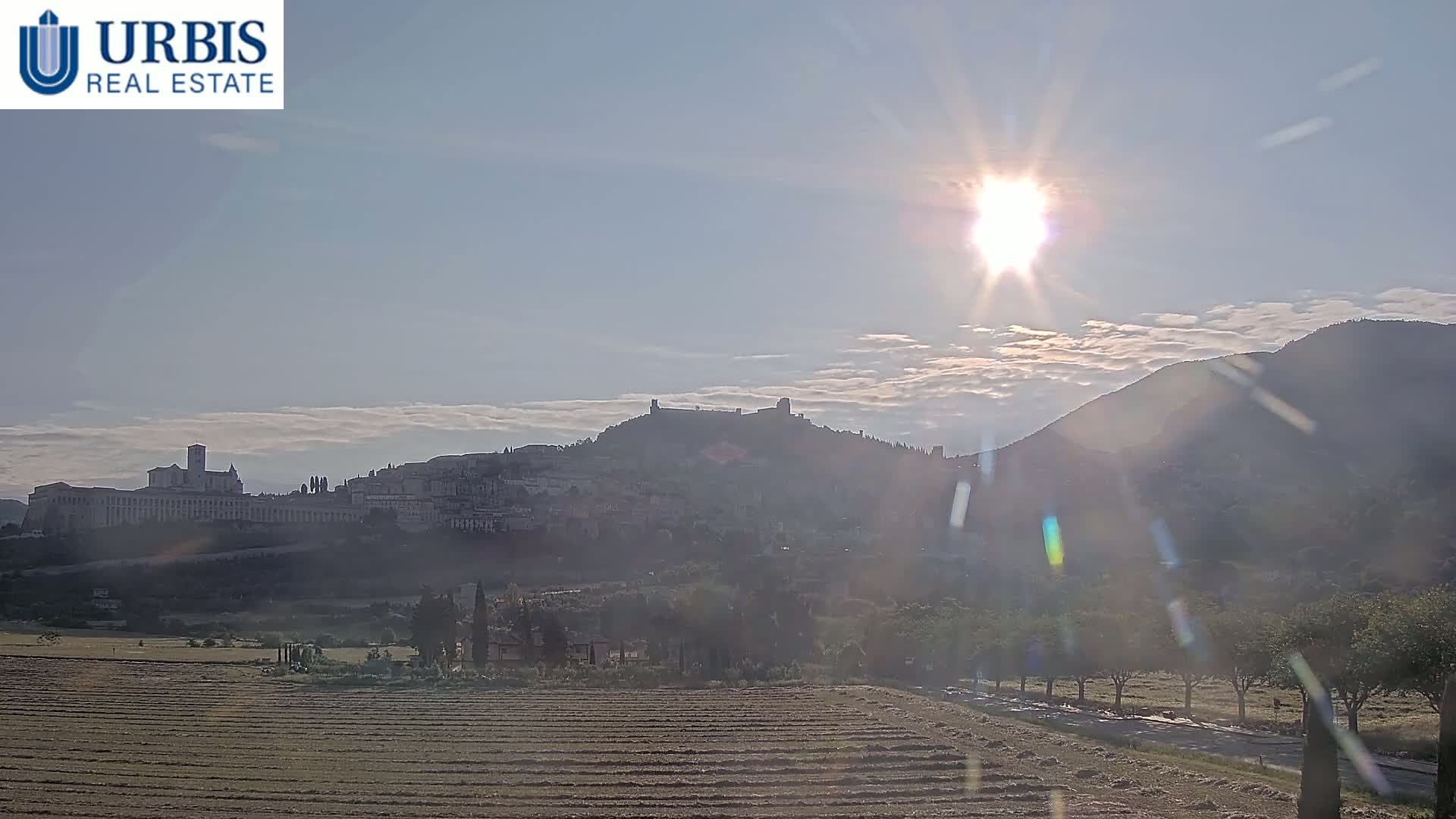A sunlit hillside town sits in the distance beyond a plowed field, under a partly cloudy sky.