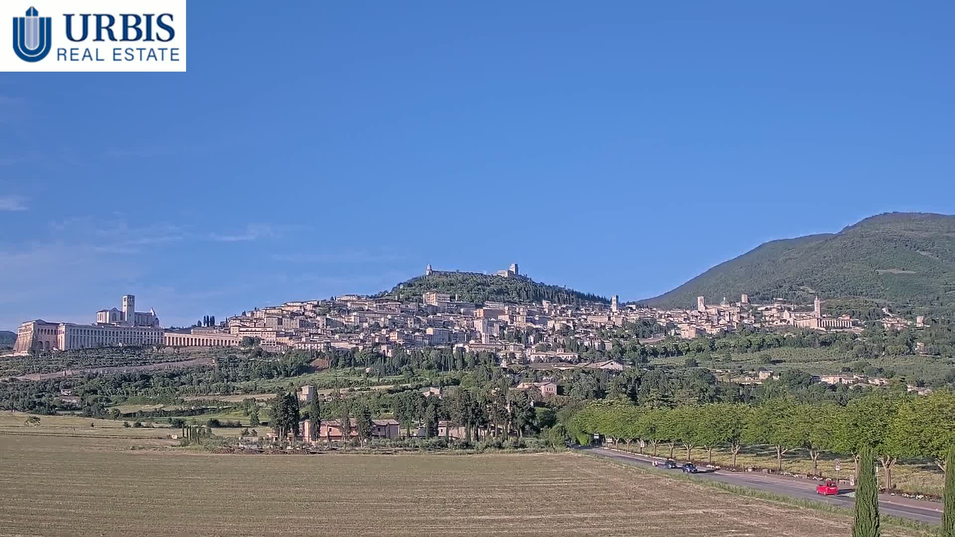 A hilltop town, nestled among green hills under a clear blue sky, is seen from a distance with a road and some cars in the foreground.
