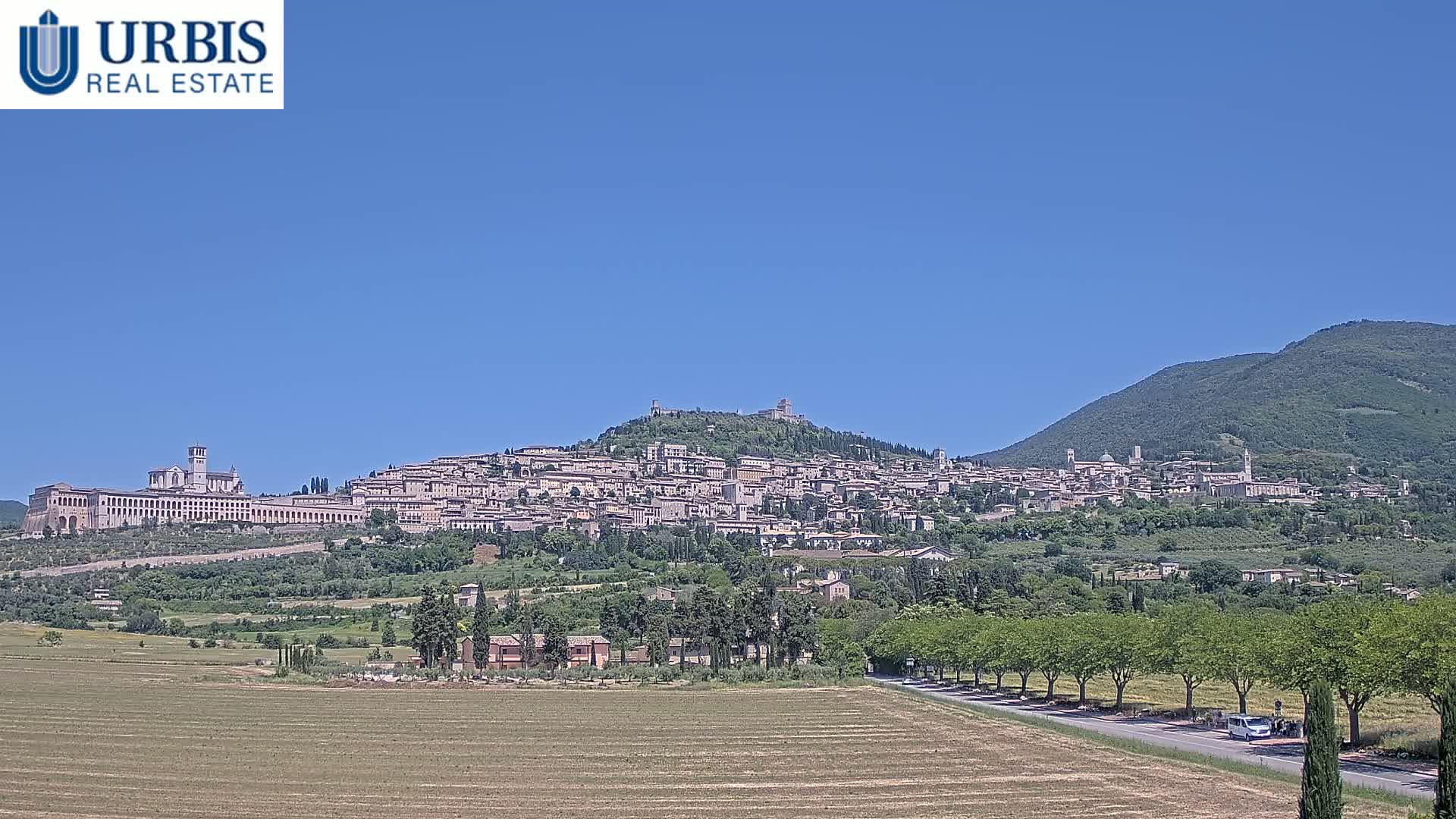 A large town sits atop a hill under a clear blue sky, with a flat expanse of land and a road in the foreground.