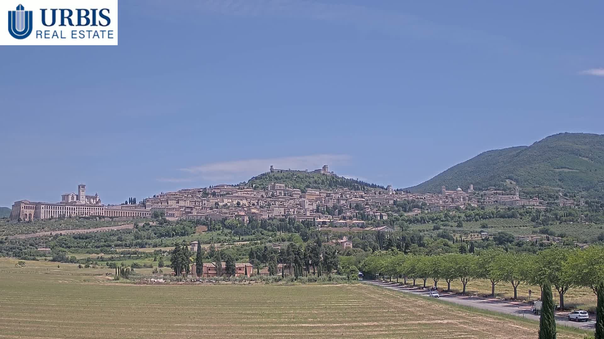 A hilltop town with numerous buildings, including a large basilica, is visible under a clear blue sky, with a flat field and tree-lined road in the foreground.