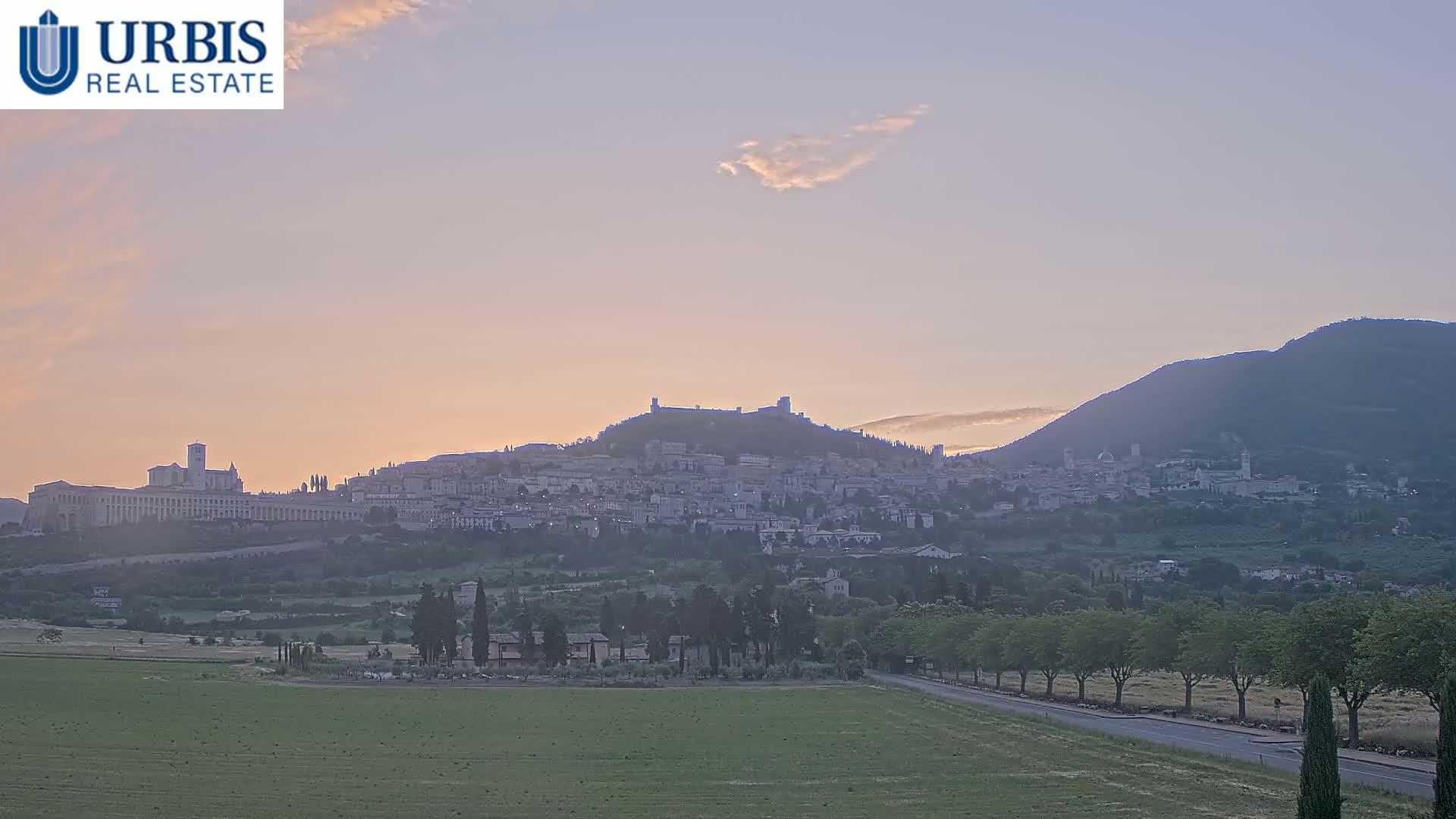 A hilltop town, silhouetted against a pale sunrise sky, is nestled between two larger hills under a hazy, calm sky.