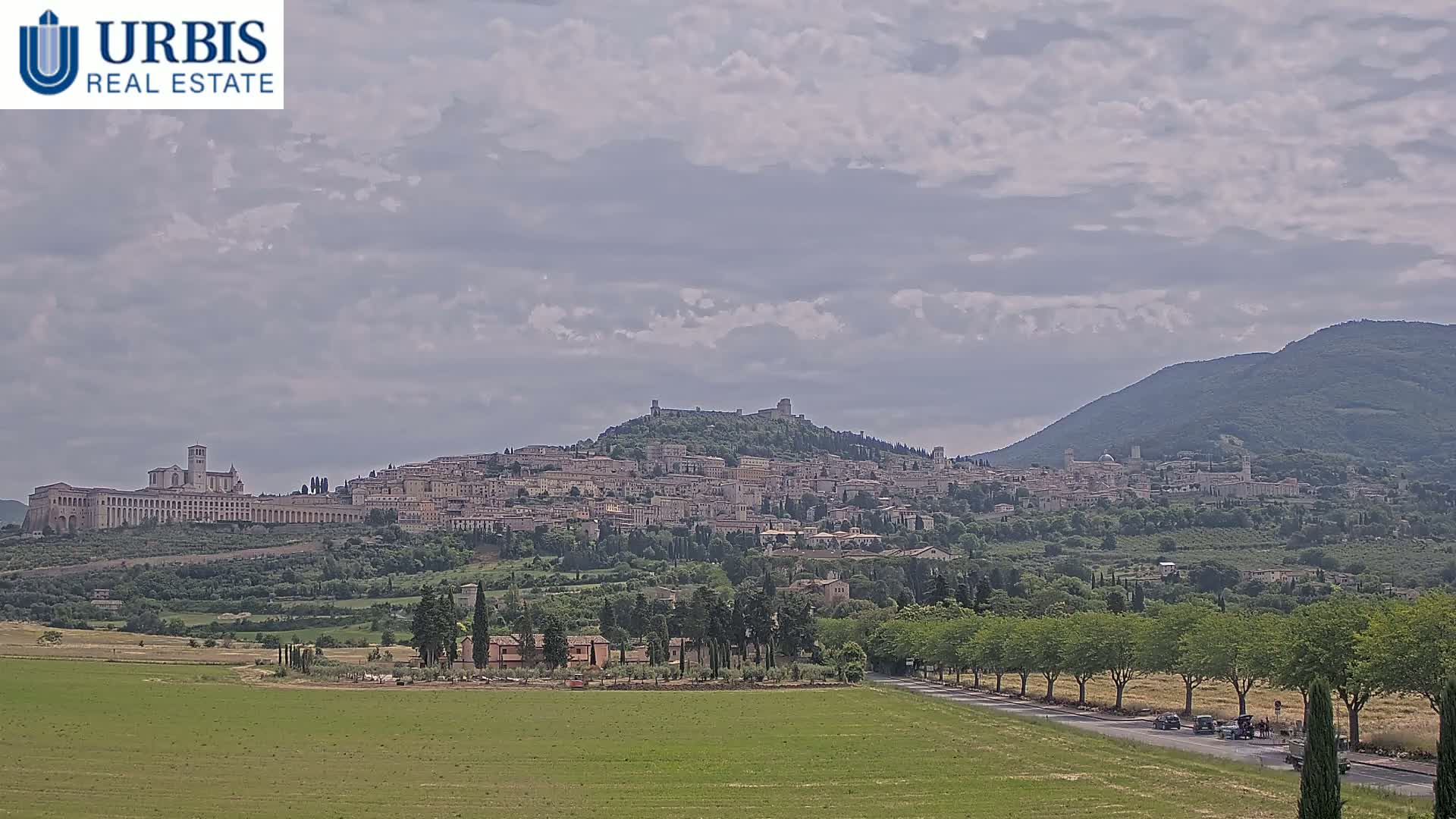A hilltop town with many buildings, including a large church, is visible under a partly cloudy sky, with a road and green fields in the foreground.